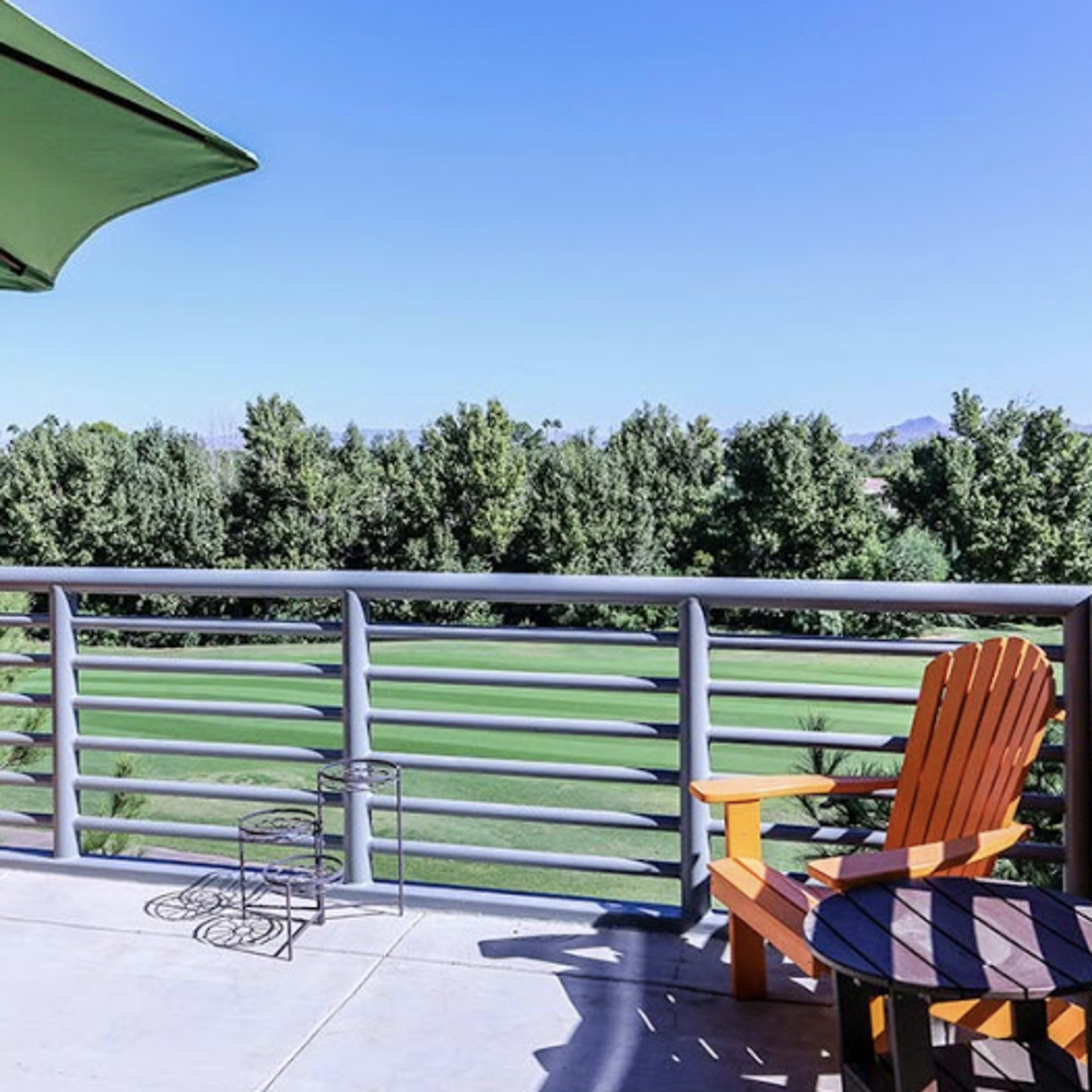 A balcony features two wooden chairs and a small table, overlooking a green landscape with trees and mountains in the distance.