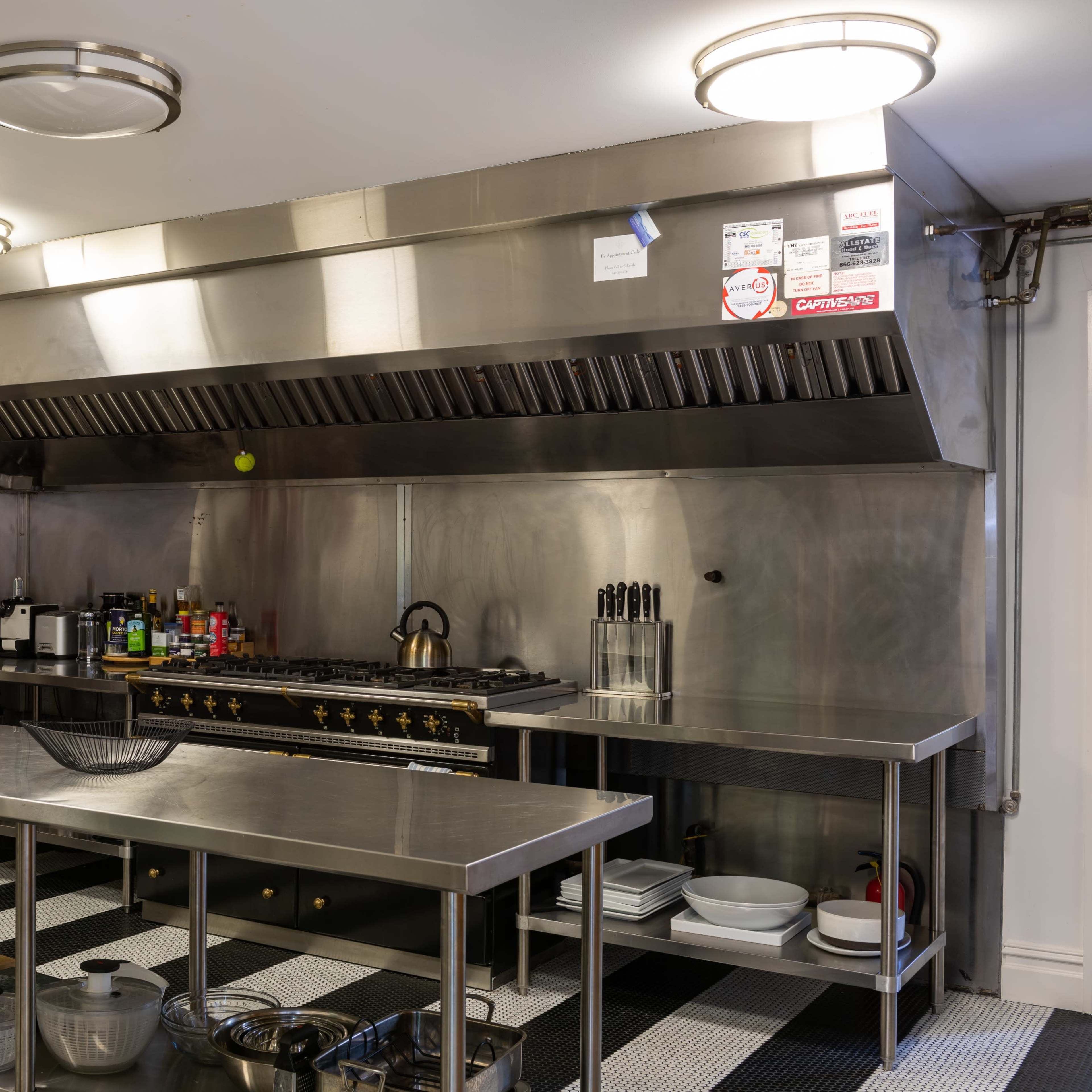 A modern commercial kitchen with stainless steel countertops, an industrial range hood, and various cooking utensils and ingredients organized on the counters.