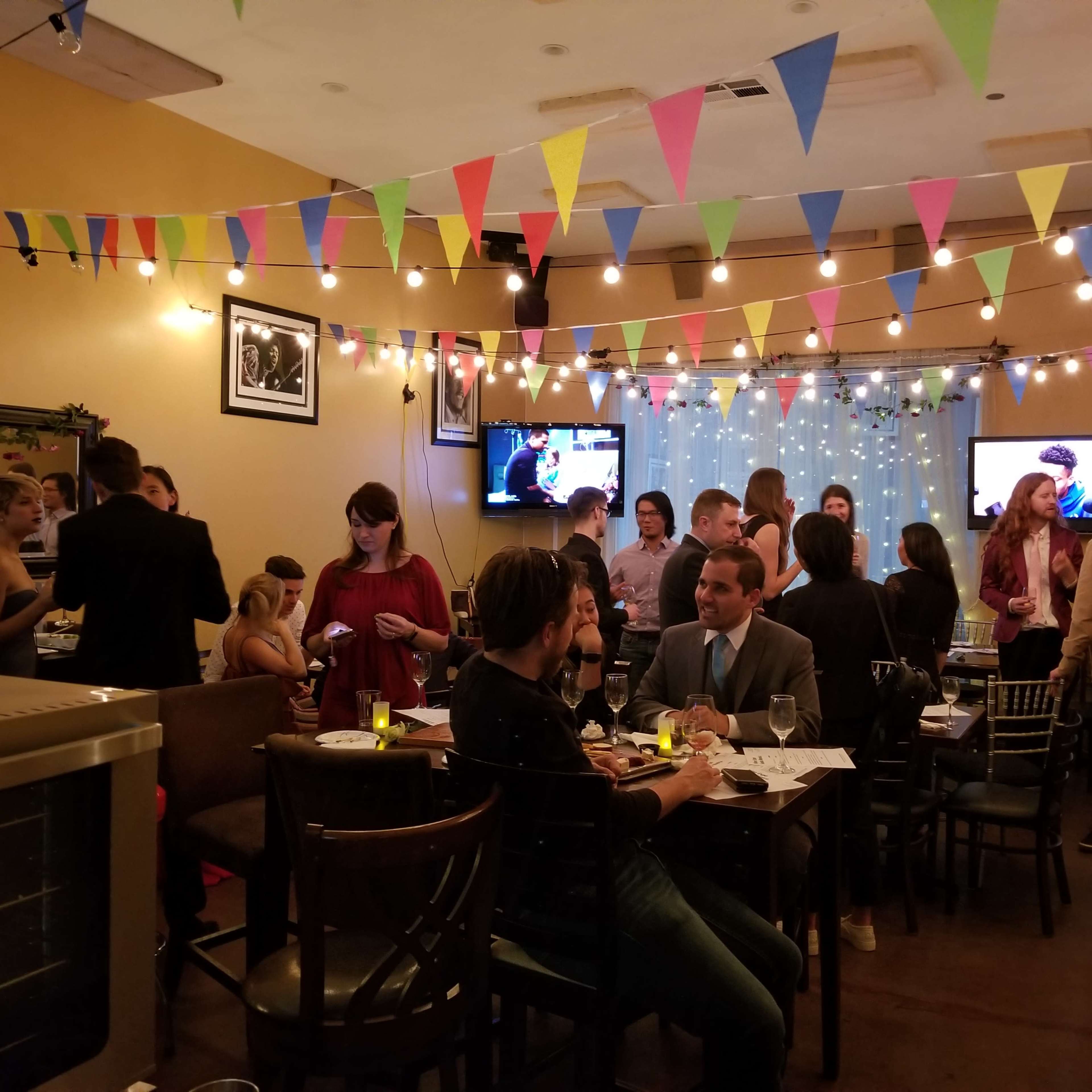 A lively social gathering takes place in a warmly lit room decorated with colorful bunting and string lights, featuring groups of people engaged in conversation.