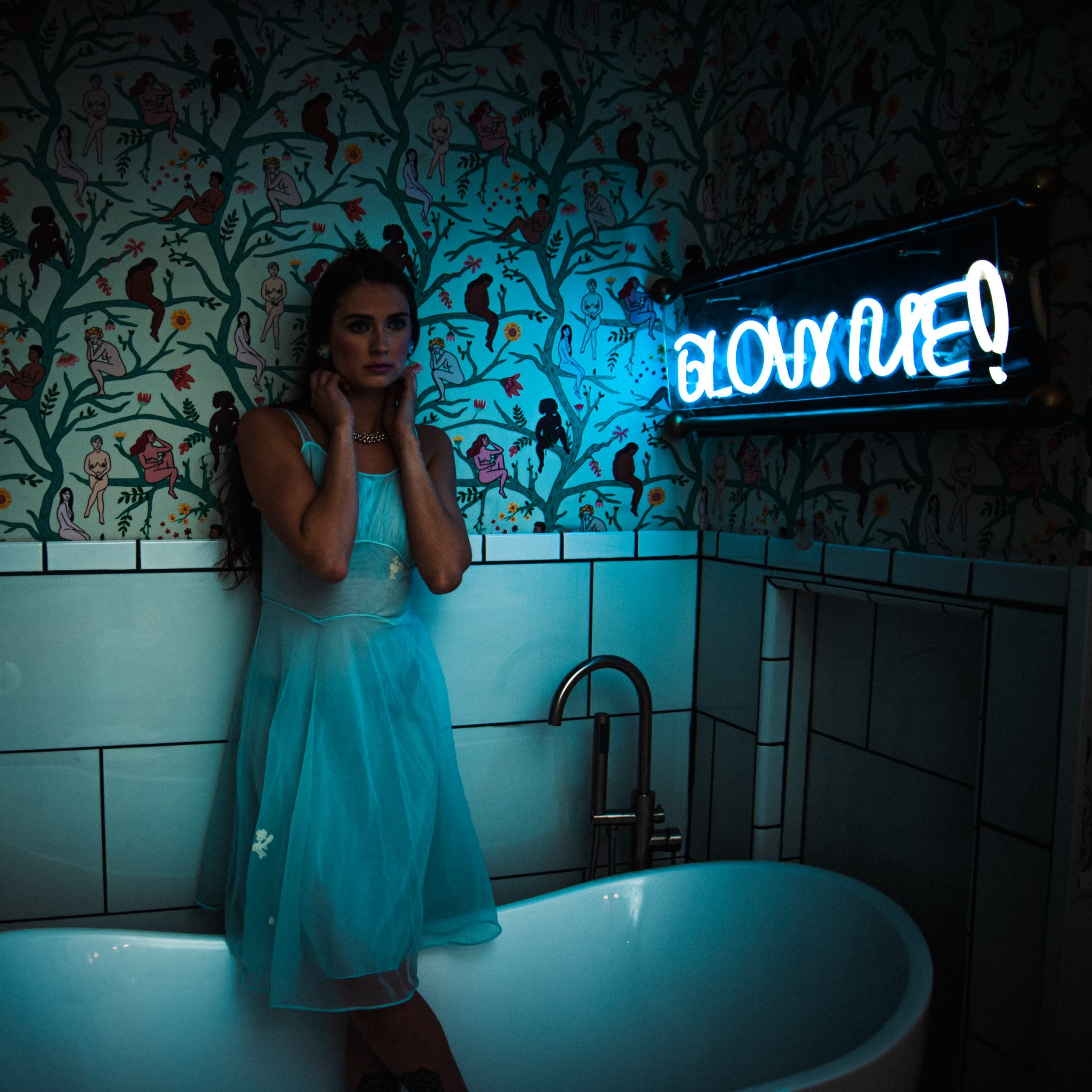 A woman in a light blue dress stands in a vintage bathtub under a neon sign that reads "GLOOMY!" in a decorated bathroom.