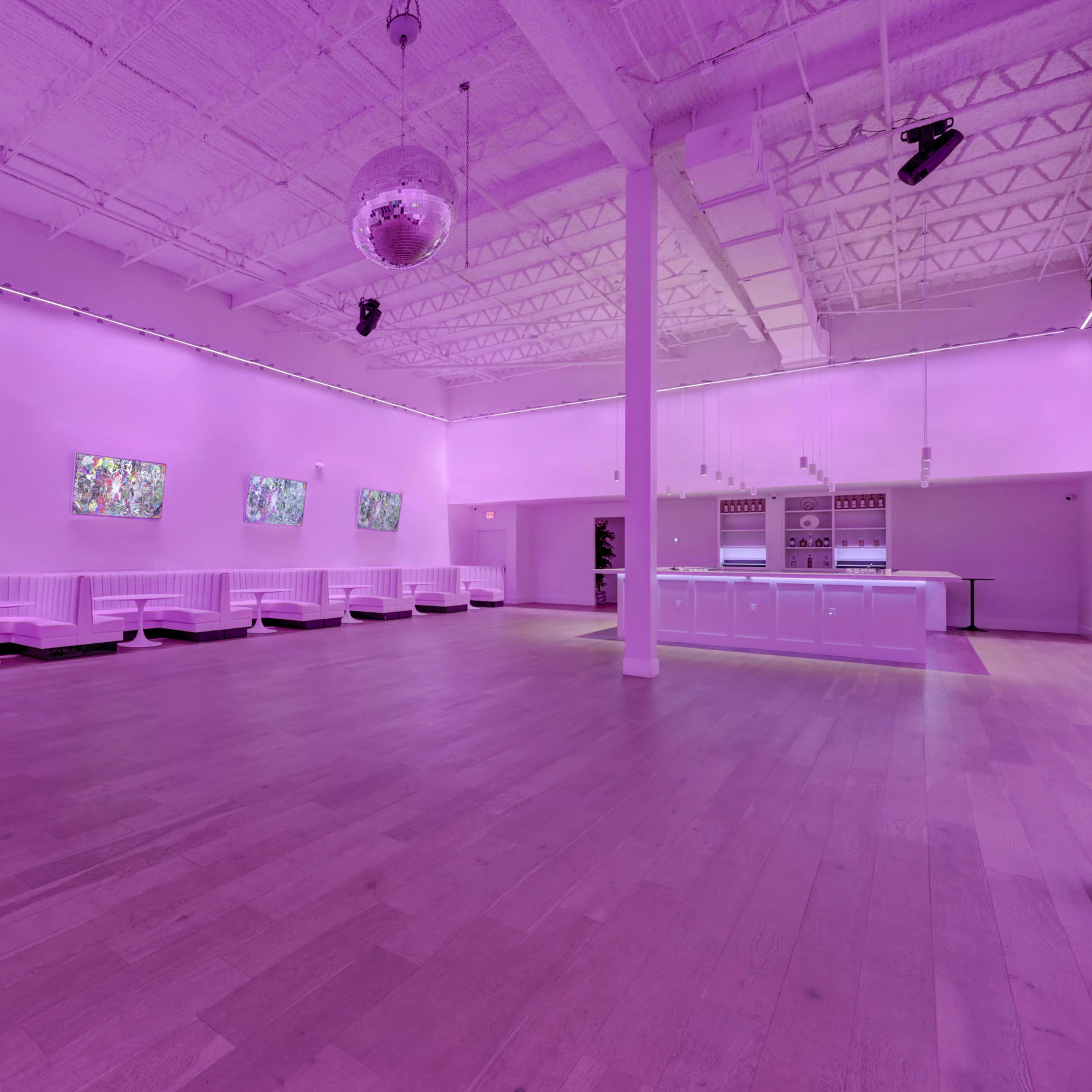 A spacious room features white benches along the walls, colorful artwork in frames, and purple lighting, with a bar area at one end.
