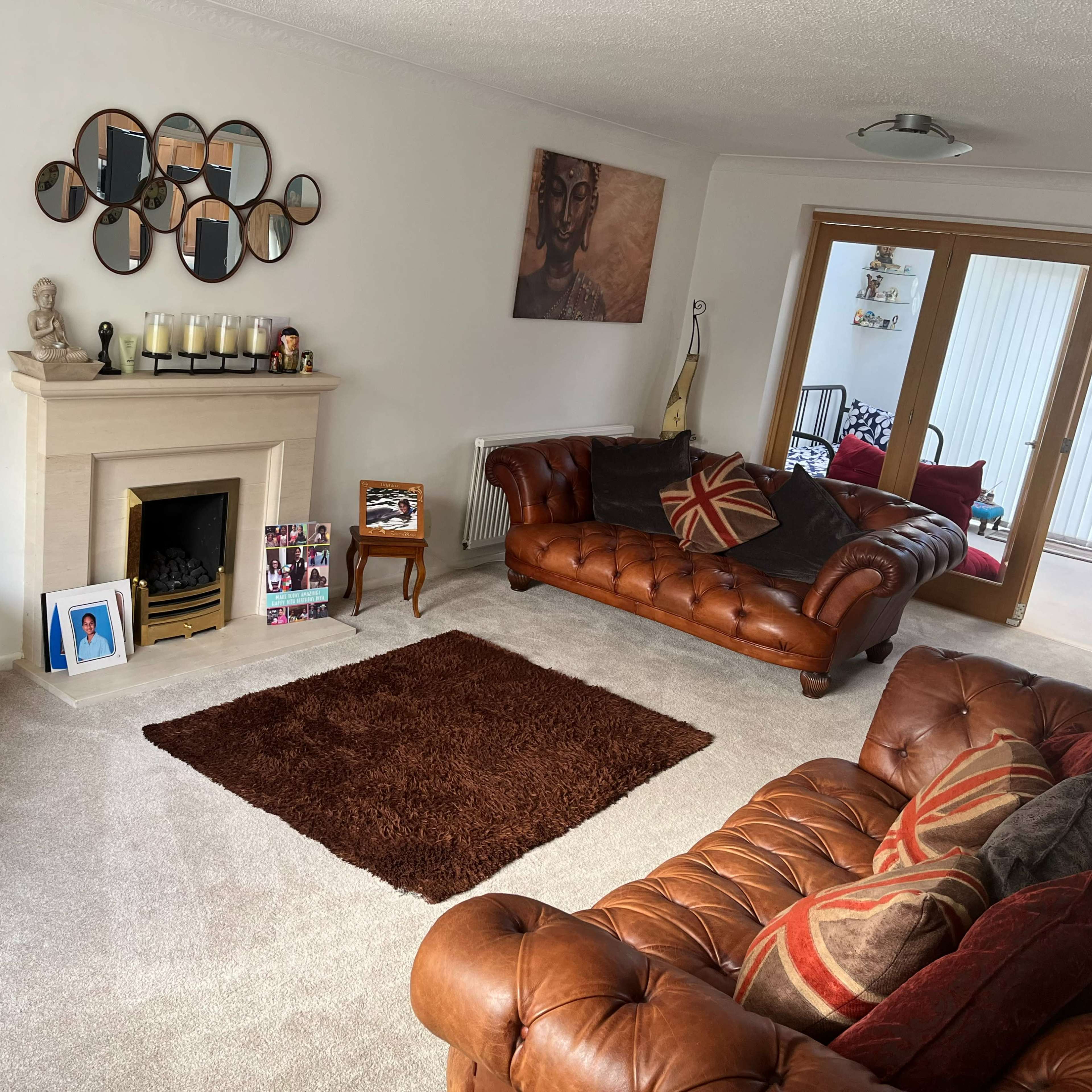 The living room features a fireplace surrounded by leather sofas, a large brown rug, and a wall adorned with round mirrors and a portrait.