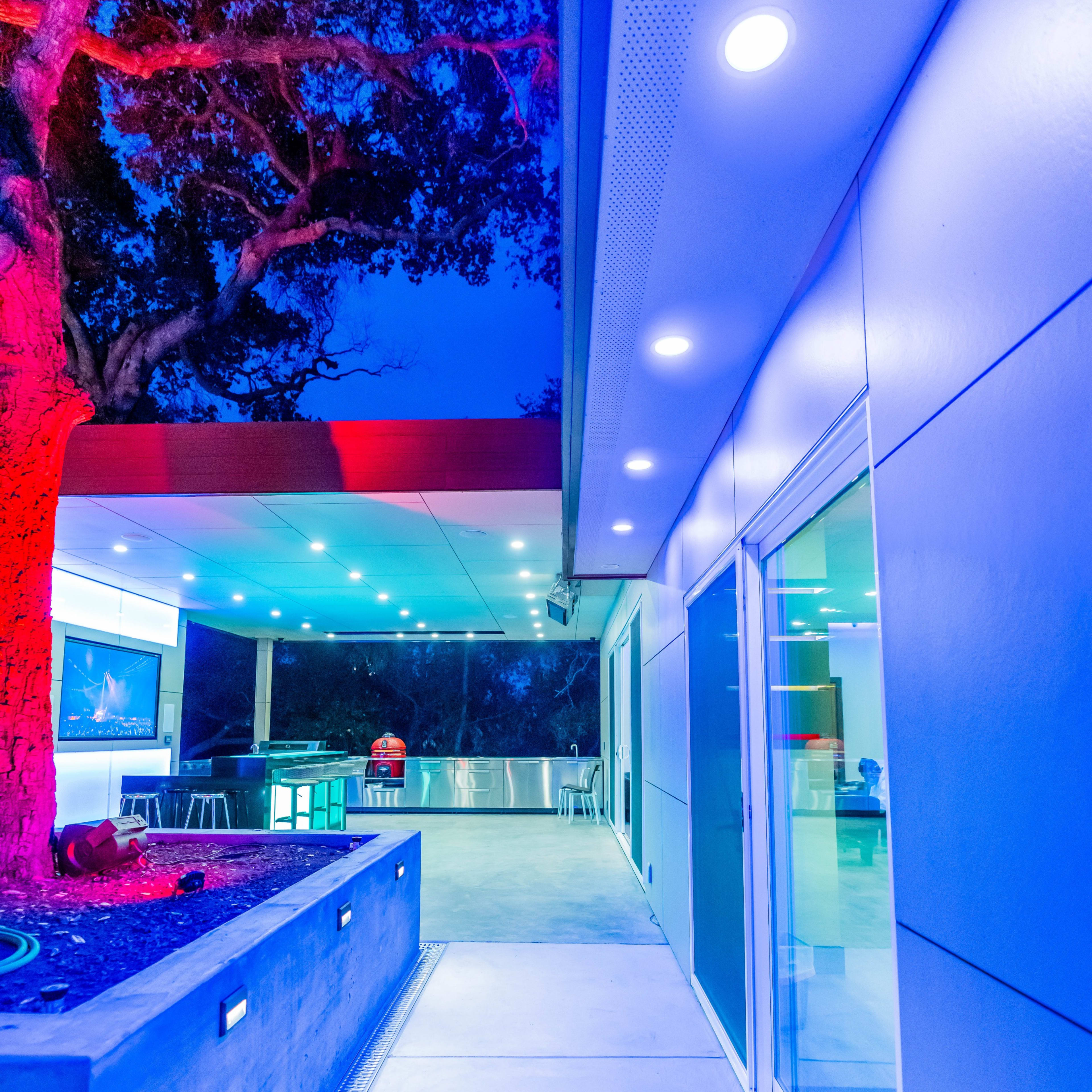 A modern, well-lit outdoor area with sleek architectural lines, featuring a pathway beside a tree and a patio space.