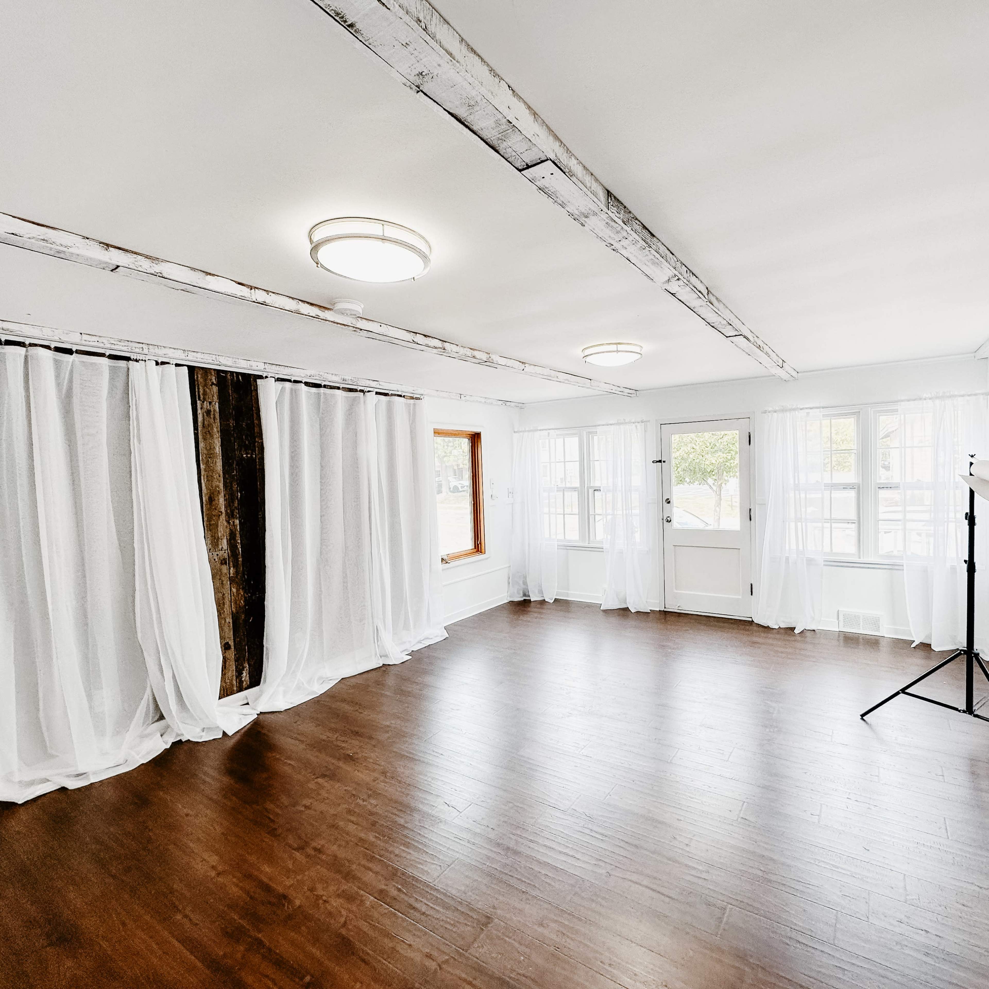 A spacious room with wooden floors, exposed beams on the ceiling, and large windows allowing natural light to enter, along with sheer curtains draped on one side.
