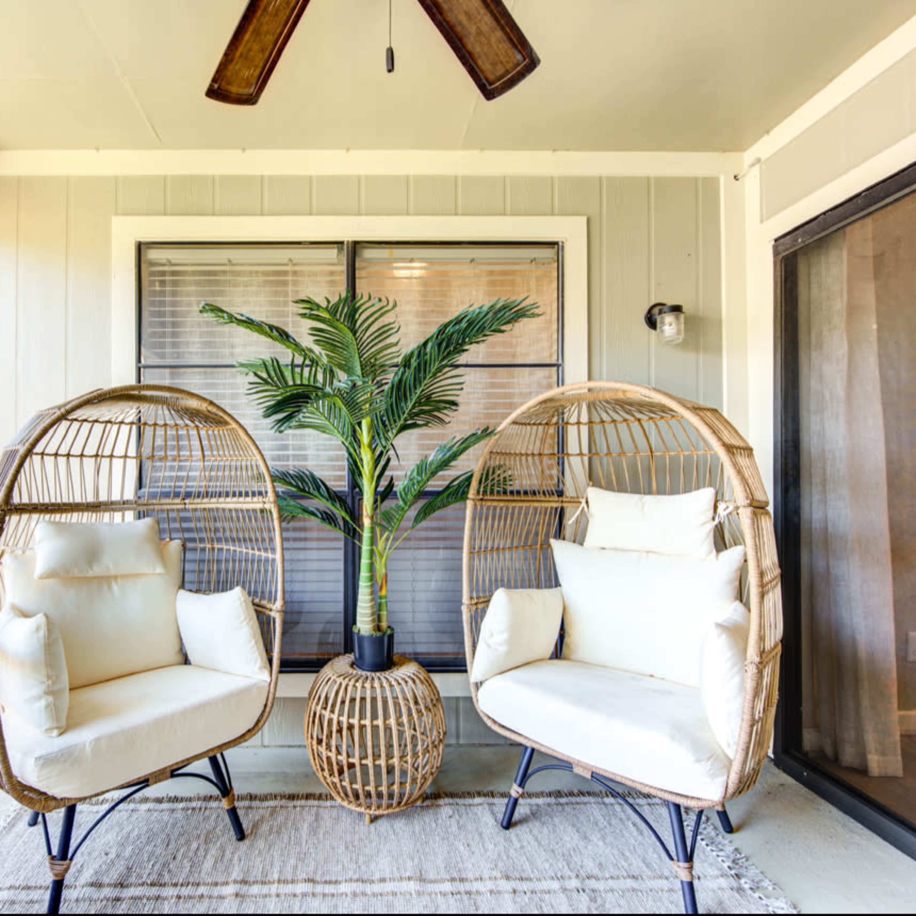 A covered balcony featuring two rattan chairs with white cushions, a potted plant between them, and a woven basket on the floor.