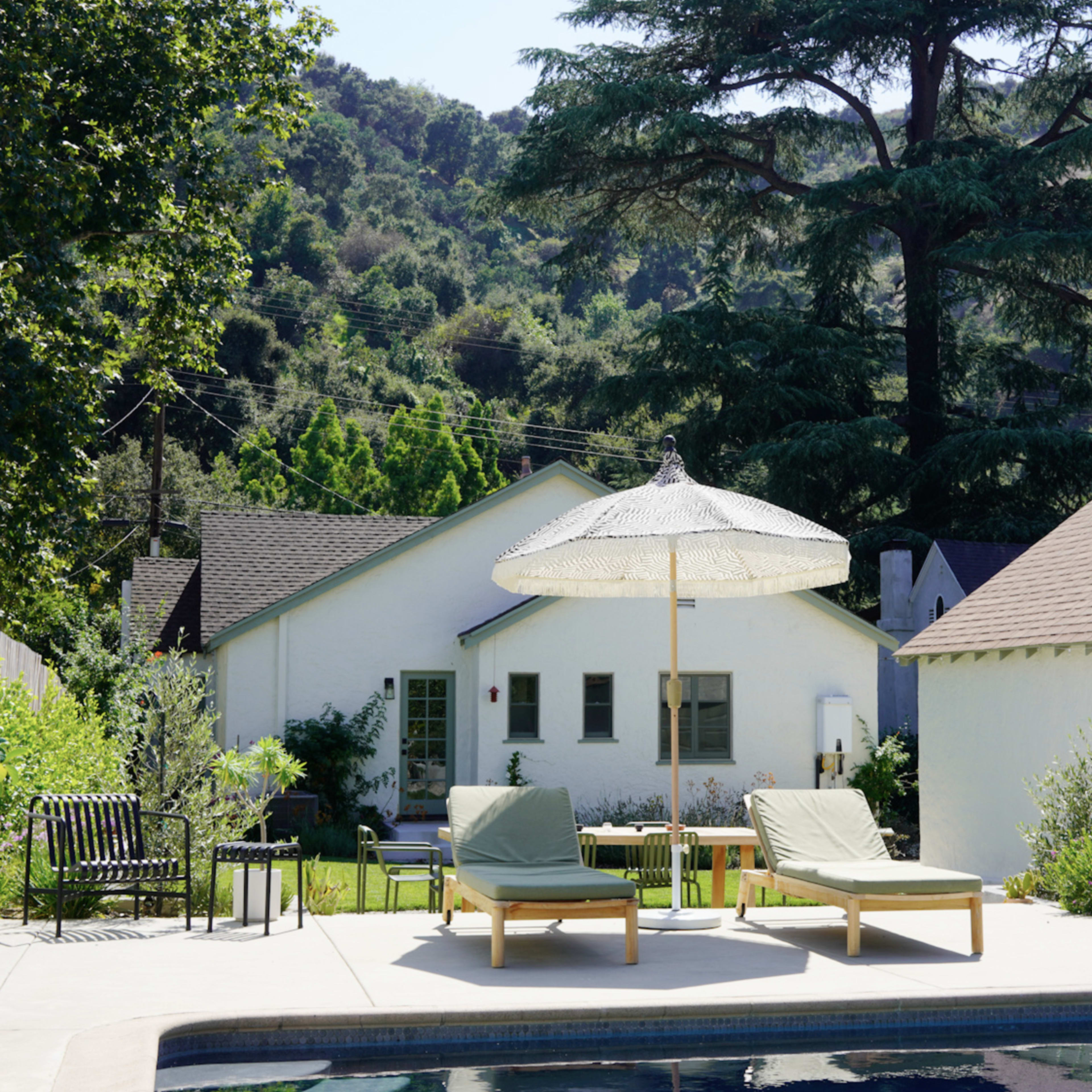 A swimming pool with two lounge chairs under a parasol is surrounded by a landscaped yard and houses against a backdrop of green hills.