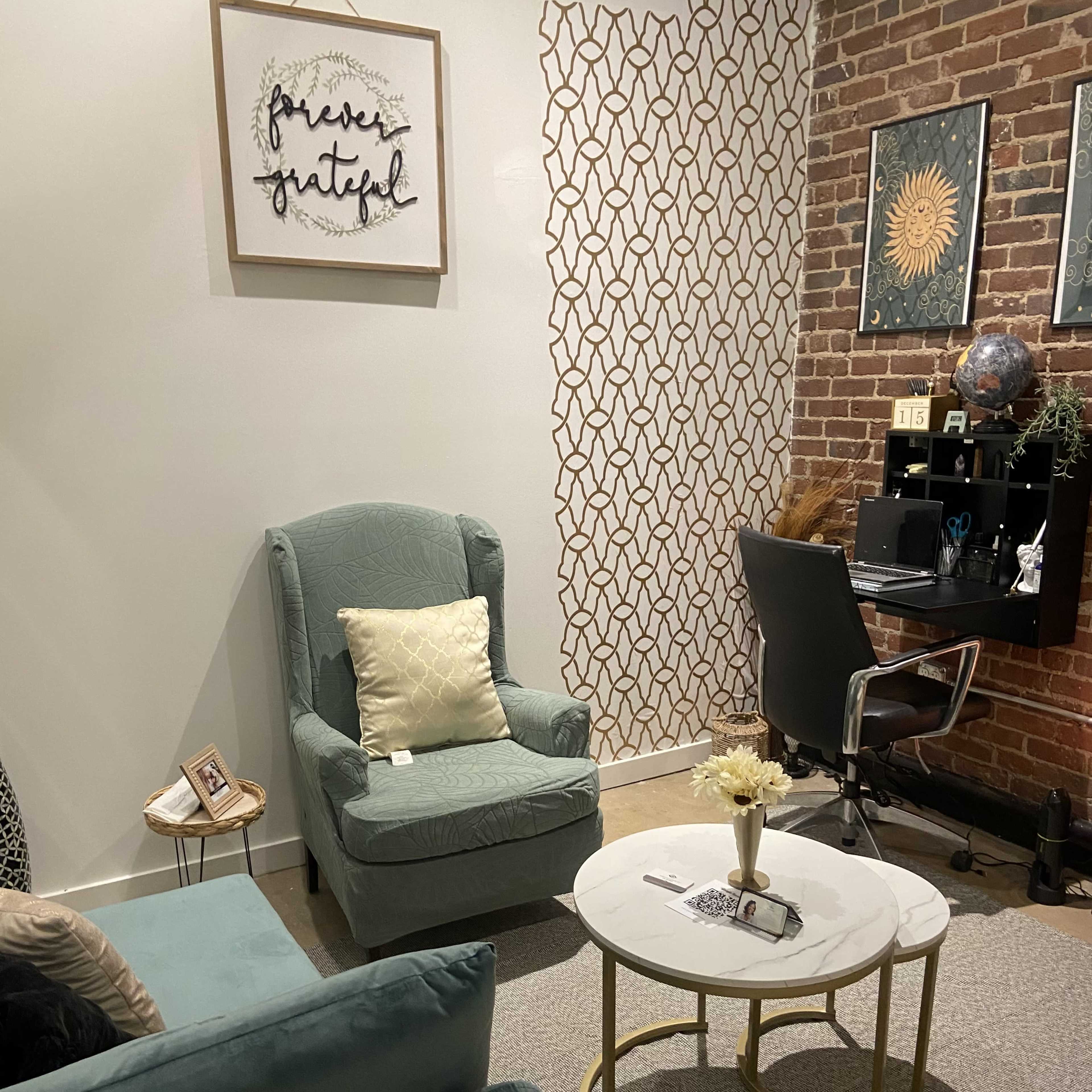 A cozy corner of a room featuring a light blue armchair, a gray circular coffee table, and a desk against a brick wall with decorative artwork.