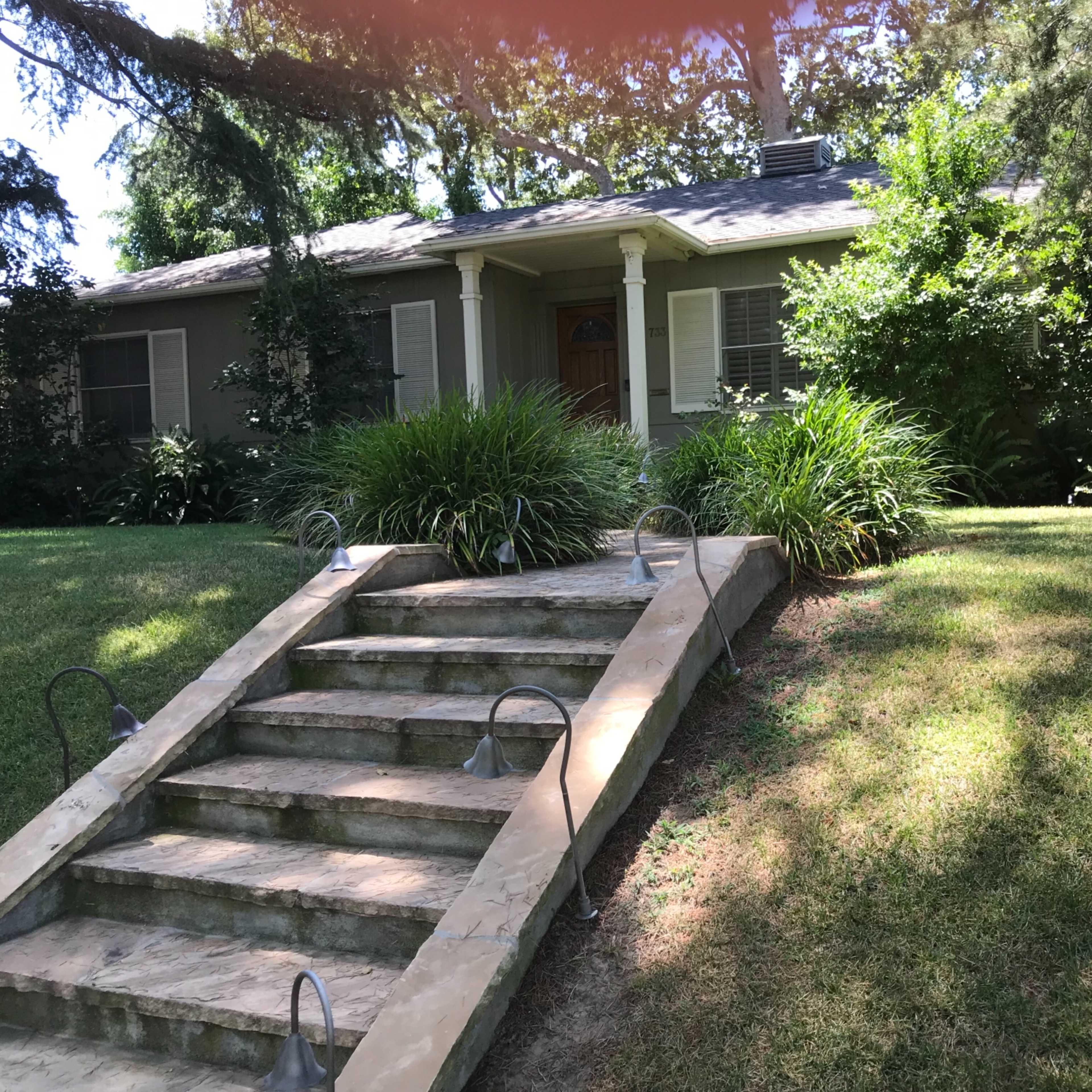 A set of stone steps leads up to a single-story, gray house surrounded by lush green foliage.