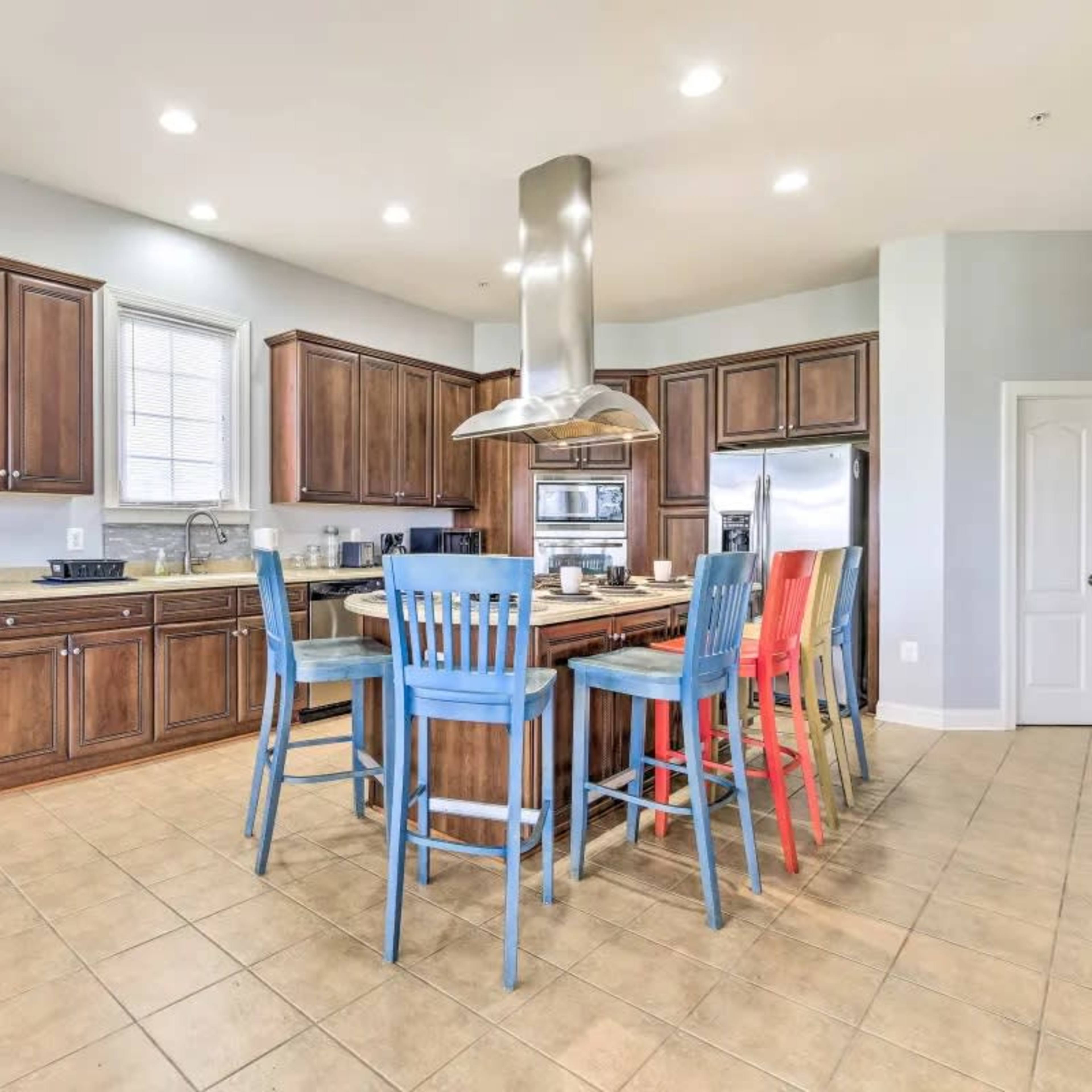 A spacious kitchen features wooden cabinets, a large island with colorful bar stools, and stainless steel appliances.