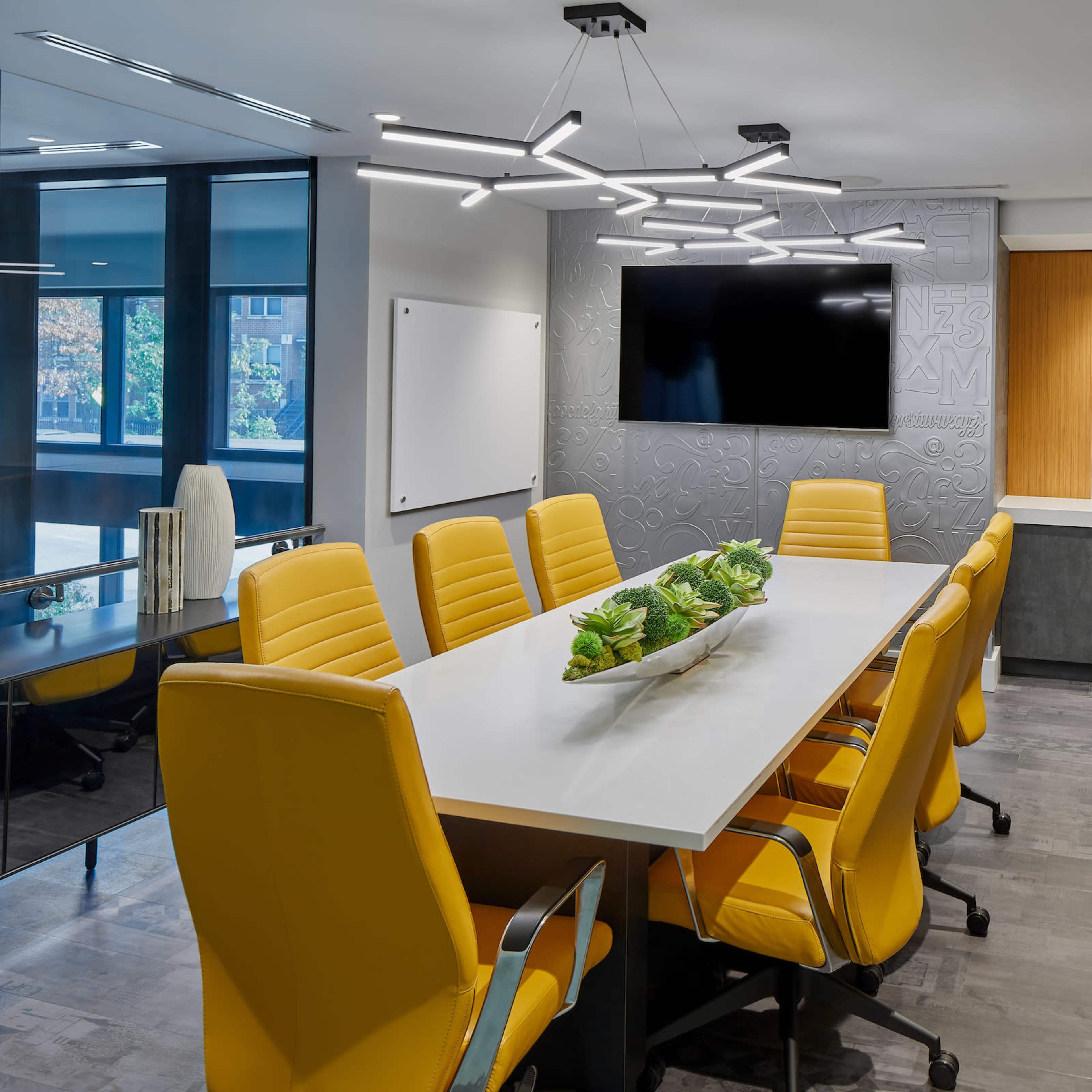 A modern conference room features a long table surrounded by yellow chairs, a wall-mounted TV, and large windows.