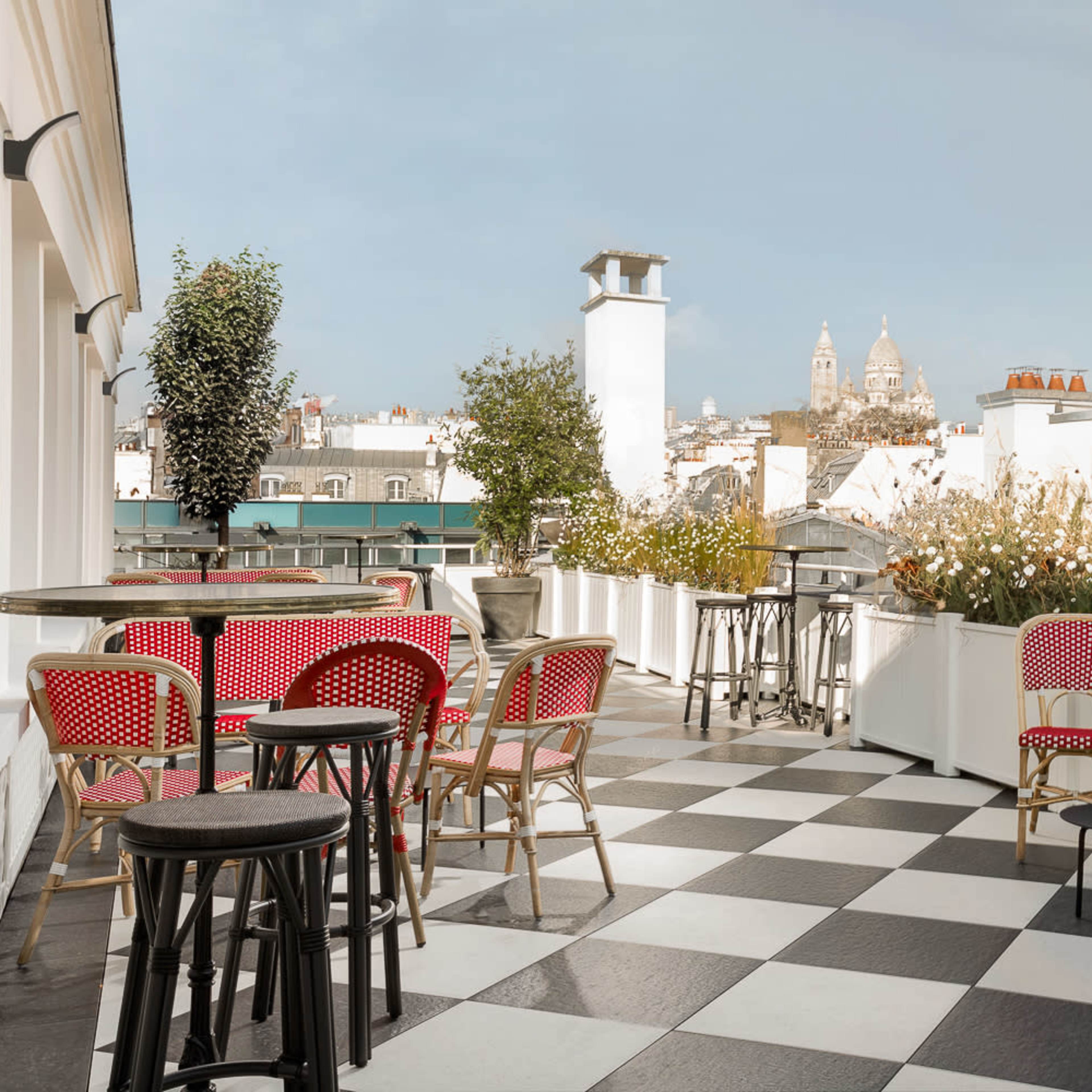 A rooftop terrace features black and white checkered flooring, surrounded by potted plants and chairs, with a distant view of a church dome.