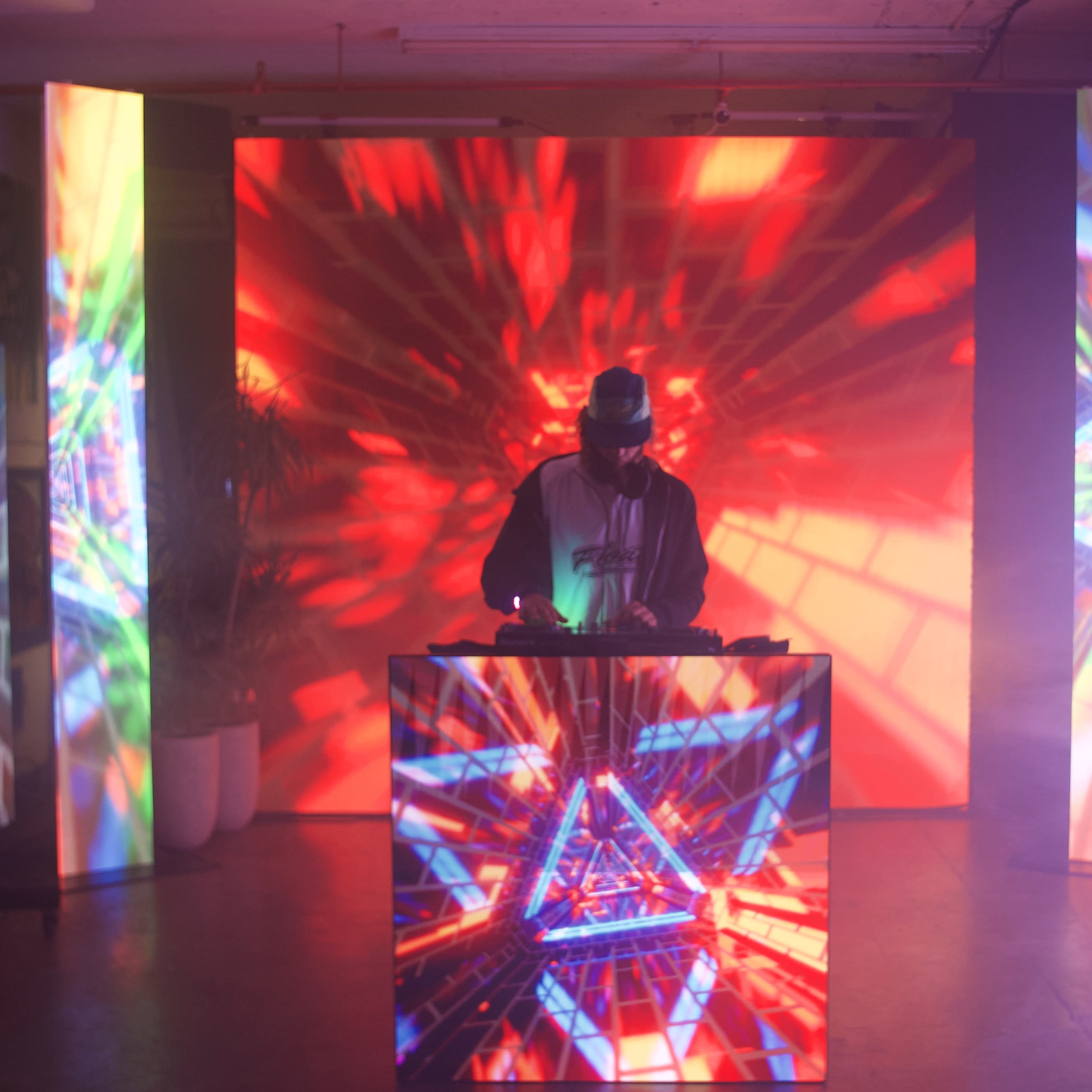 A DJ stands behind a console in a colorful, illuminated room with large screens displaying vibrant, abstract graphics.