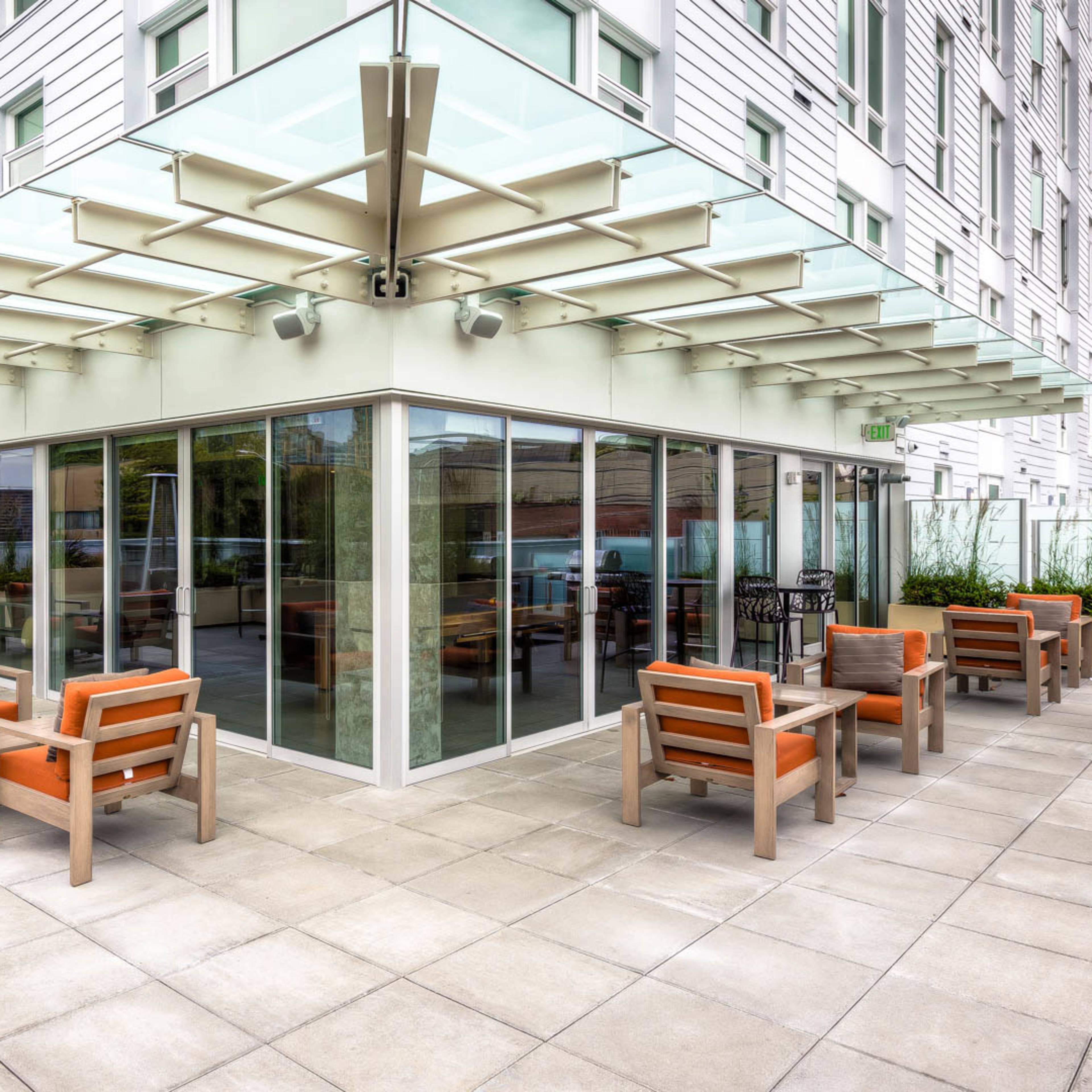 A modern outdoor seating area with metal and wooden furniture, surrounded by glass walls and planters.