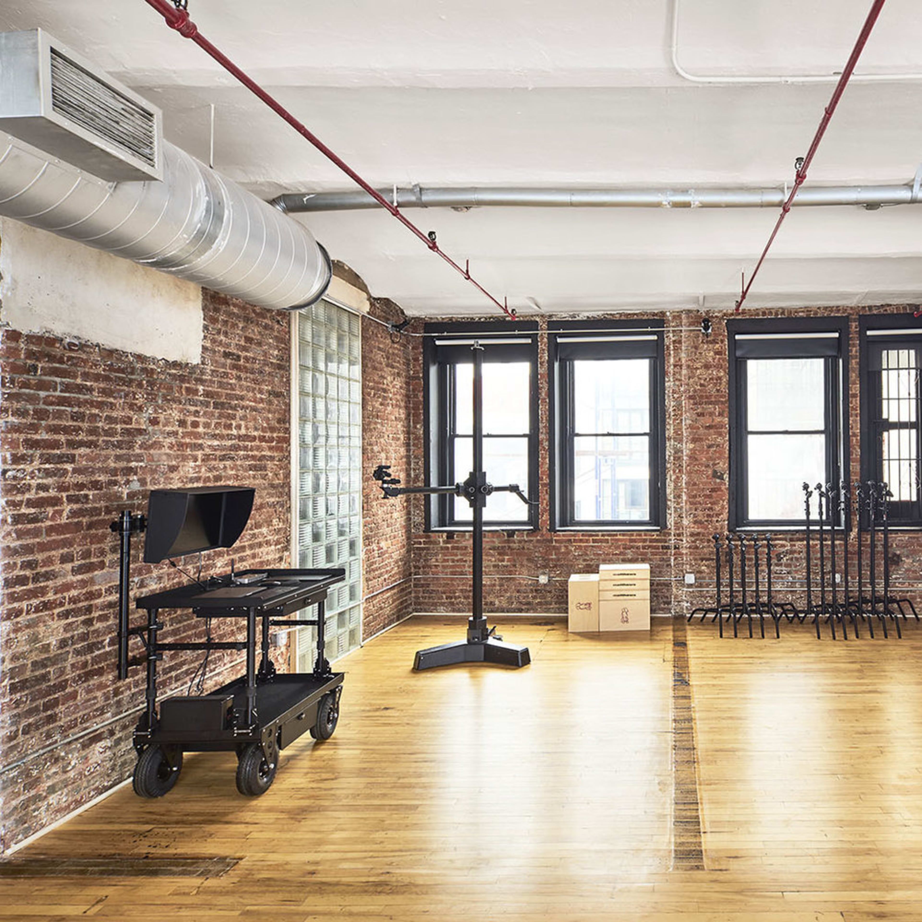 A spacious room with exposed brick walls, hardwood flooring, and large windows, featuring a black cart with monitors and several metal stands in the corner.