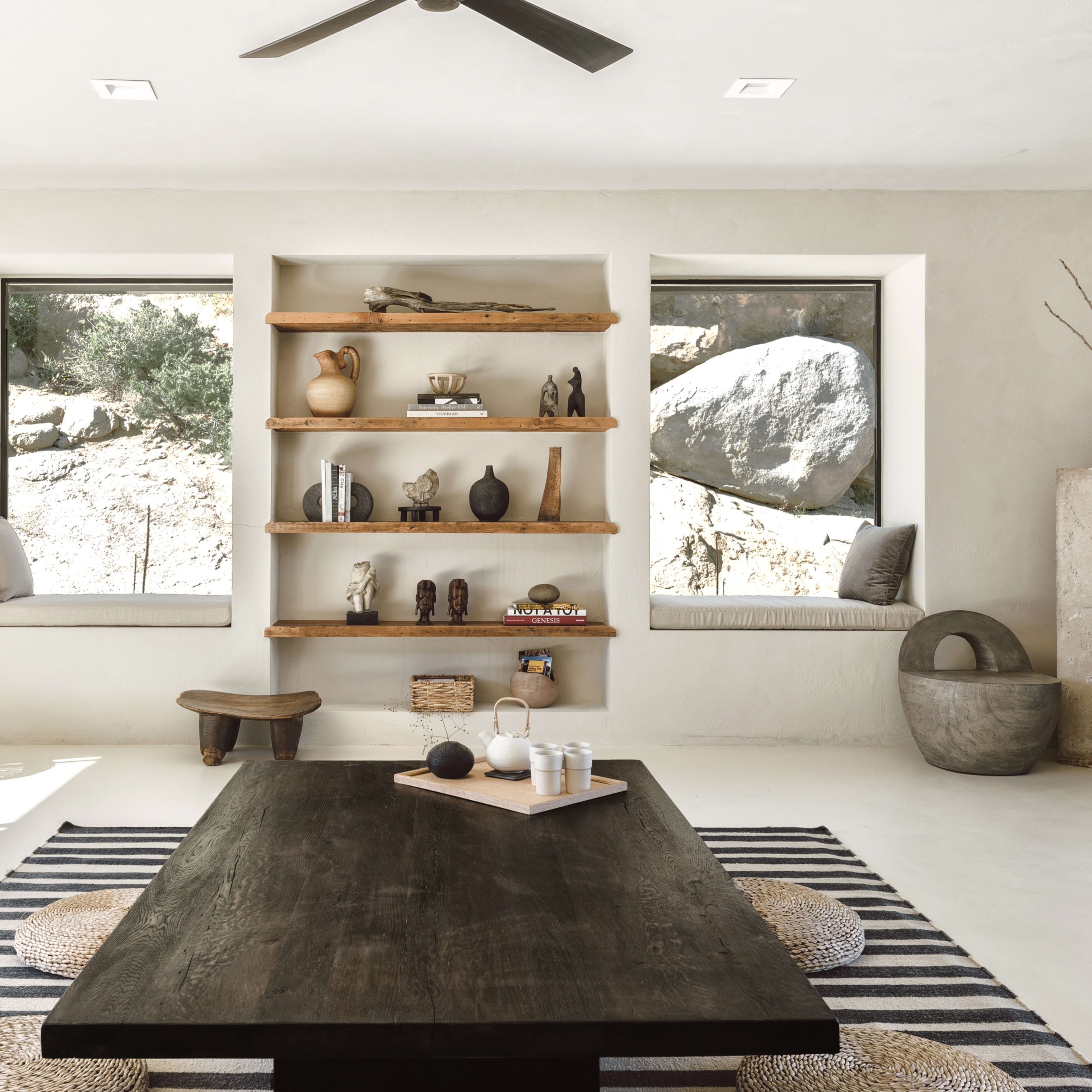 A minimalist room with a large dark wooden table, a striped rug, and shelves displaying various decorative objects, flanked by two windows with window seats.