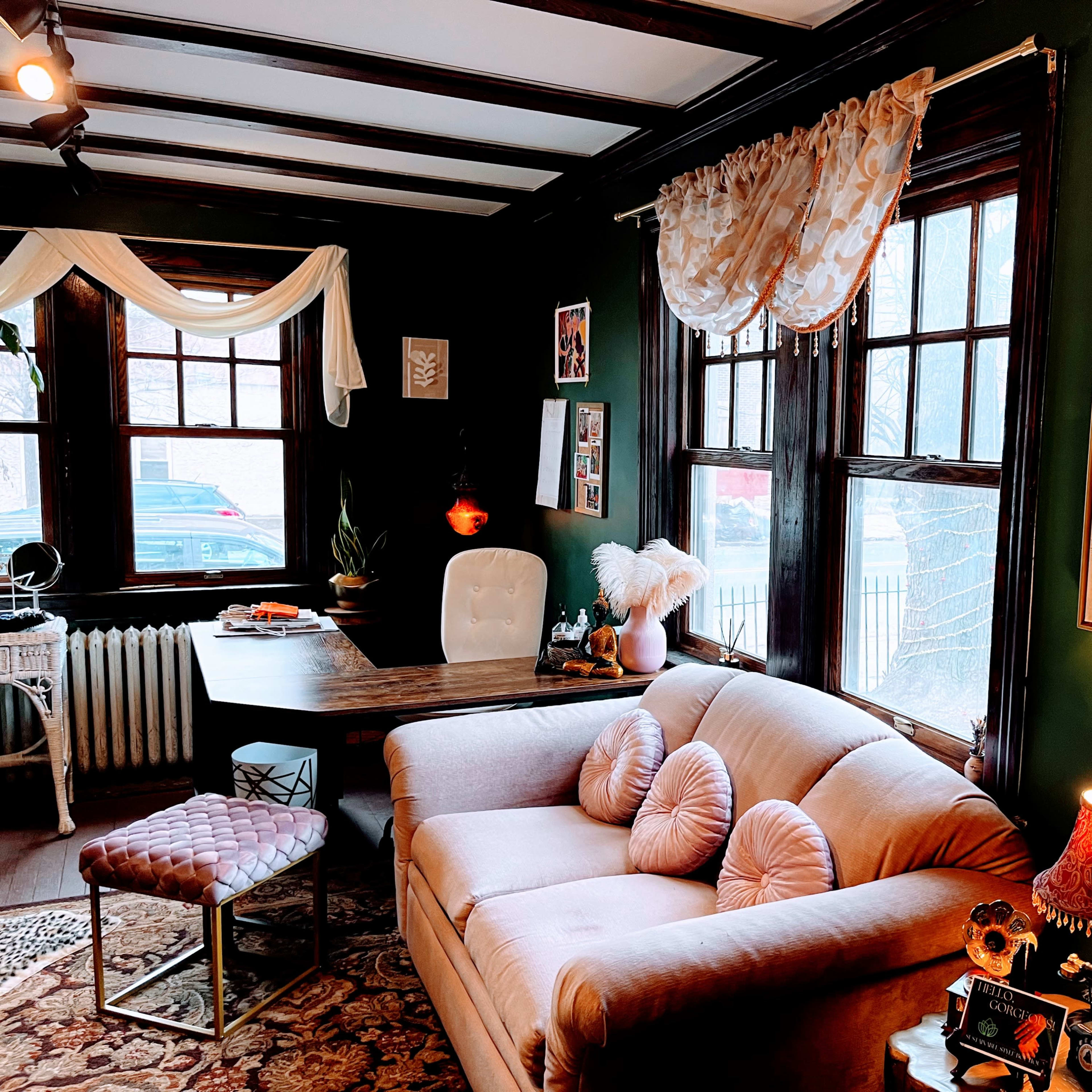 A cozy living room with a pink sofa, a wooden table, and several windows, decorated with plants and framed photographs.
