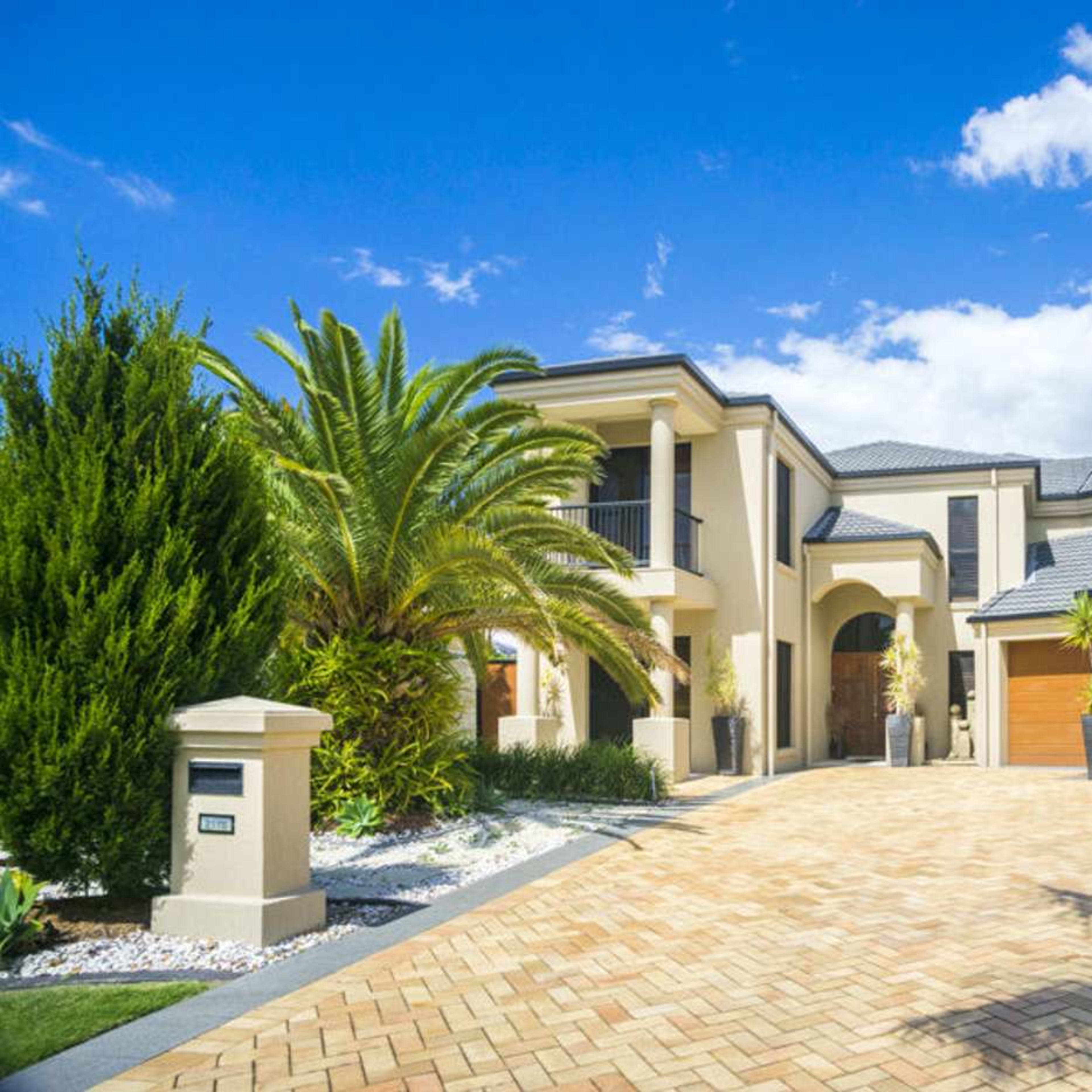 A large, modern two-story house with a paved driveway and palm trees in the front yard.
