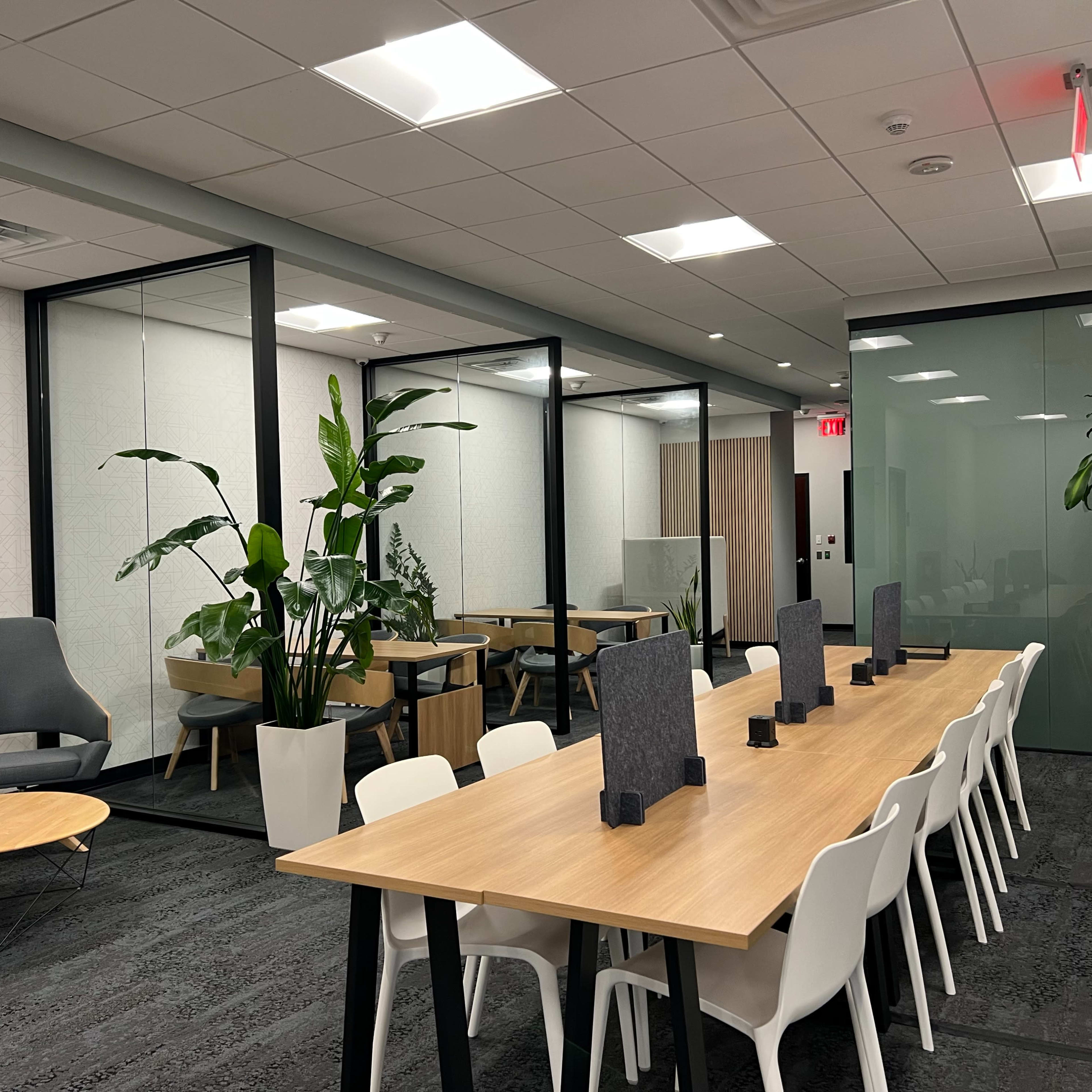 A modern office space featuring a long wooden table surrounded by white chairs, potted plants, and glass partitioned meeting rooms.