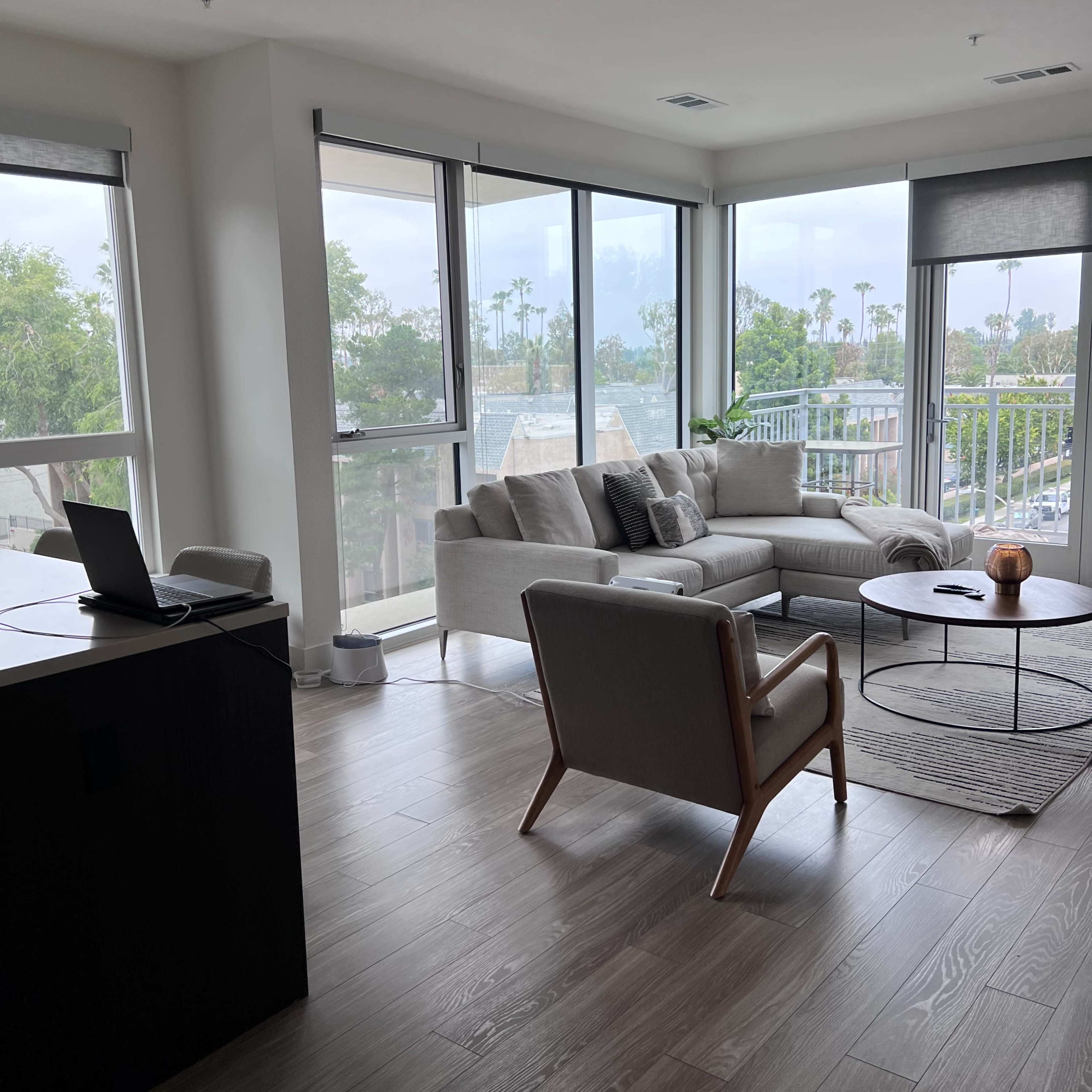 A modern living room features a light-colored sofa, a round coffee table, and large windows overlooking greenery and palm trees.