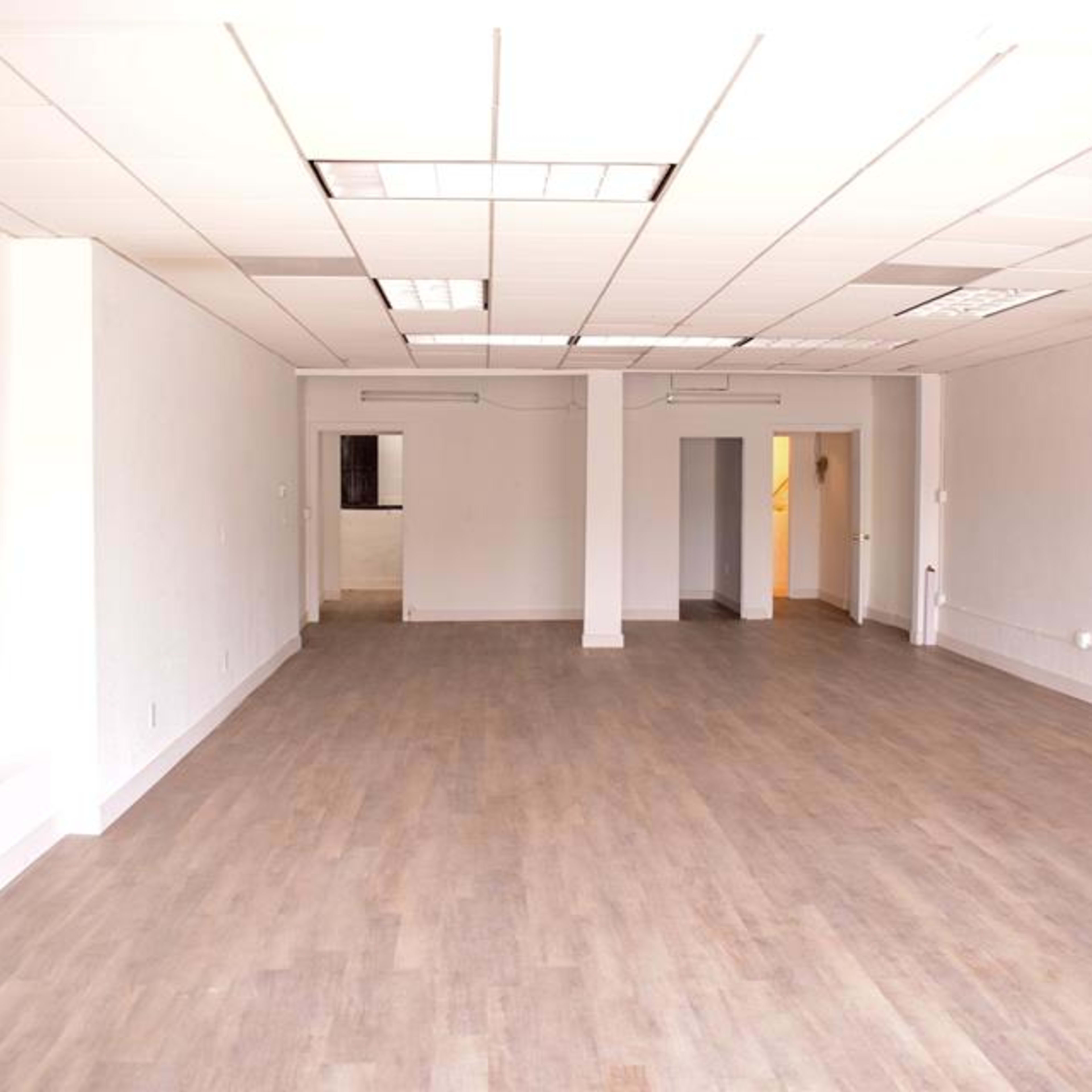 A spacious, empty room with wood-like flooring and white walls, featuring multiple doorways and fluorescent lighting.