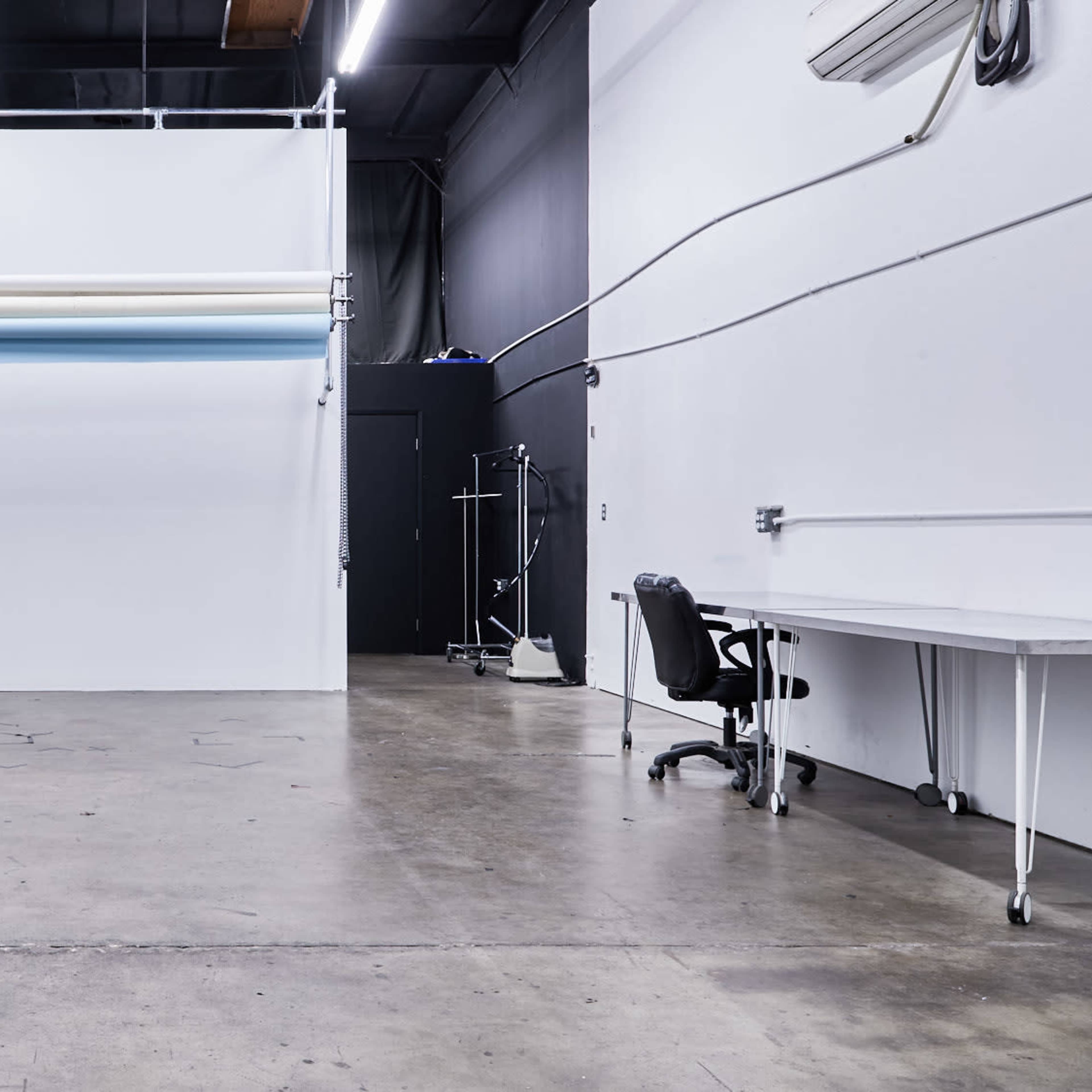 A spacious studio with a concrete floor, bare white walls, and a large backdrop holder mounted on the wall, alongside a desk and a chair.
