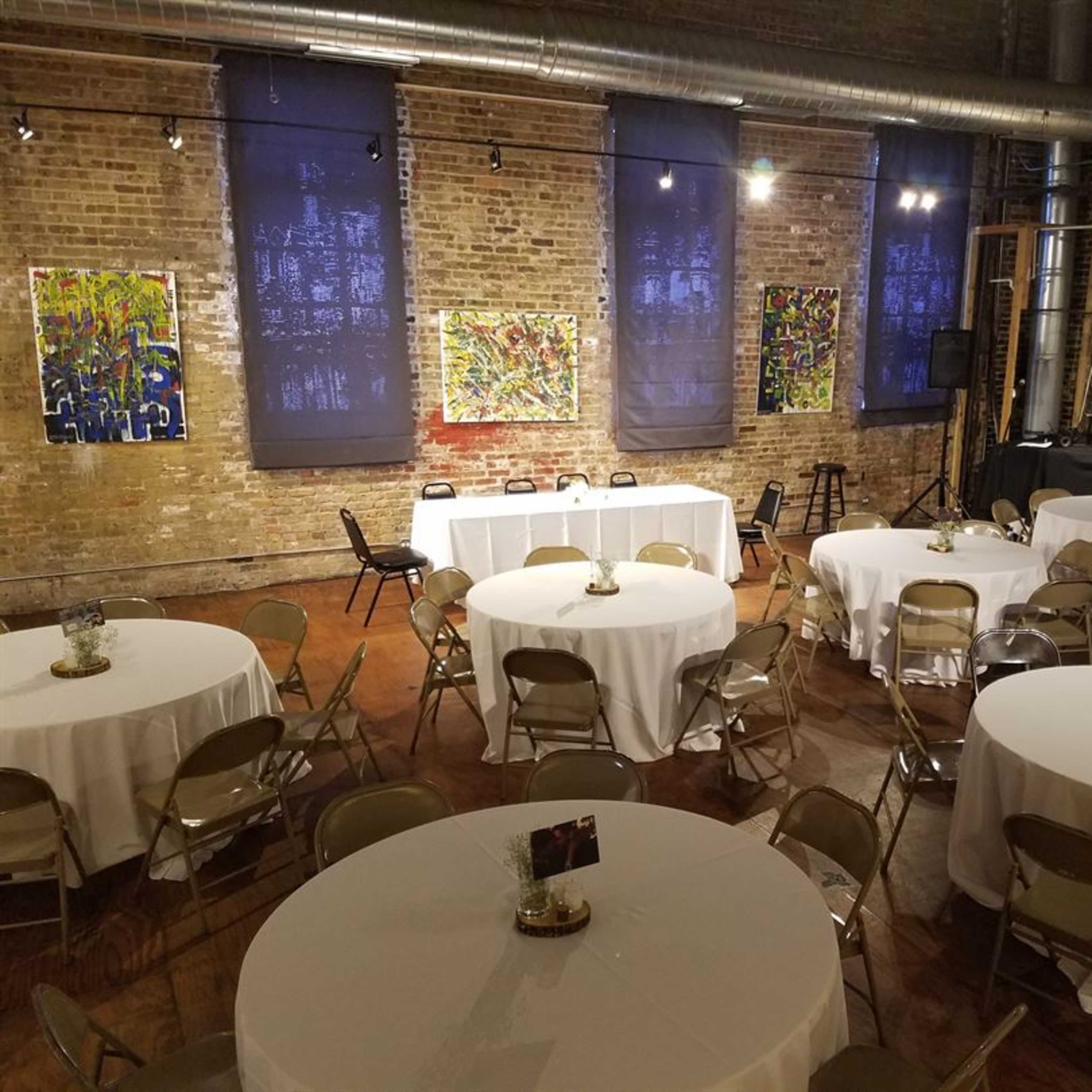 A large indoor event space with round tables covered in white tablecloths, surrounded by brick walls adorned with colorful artwork.