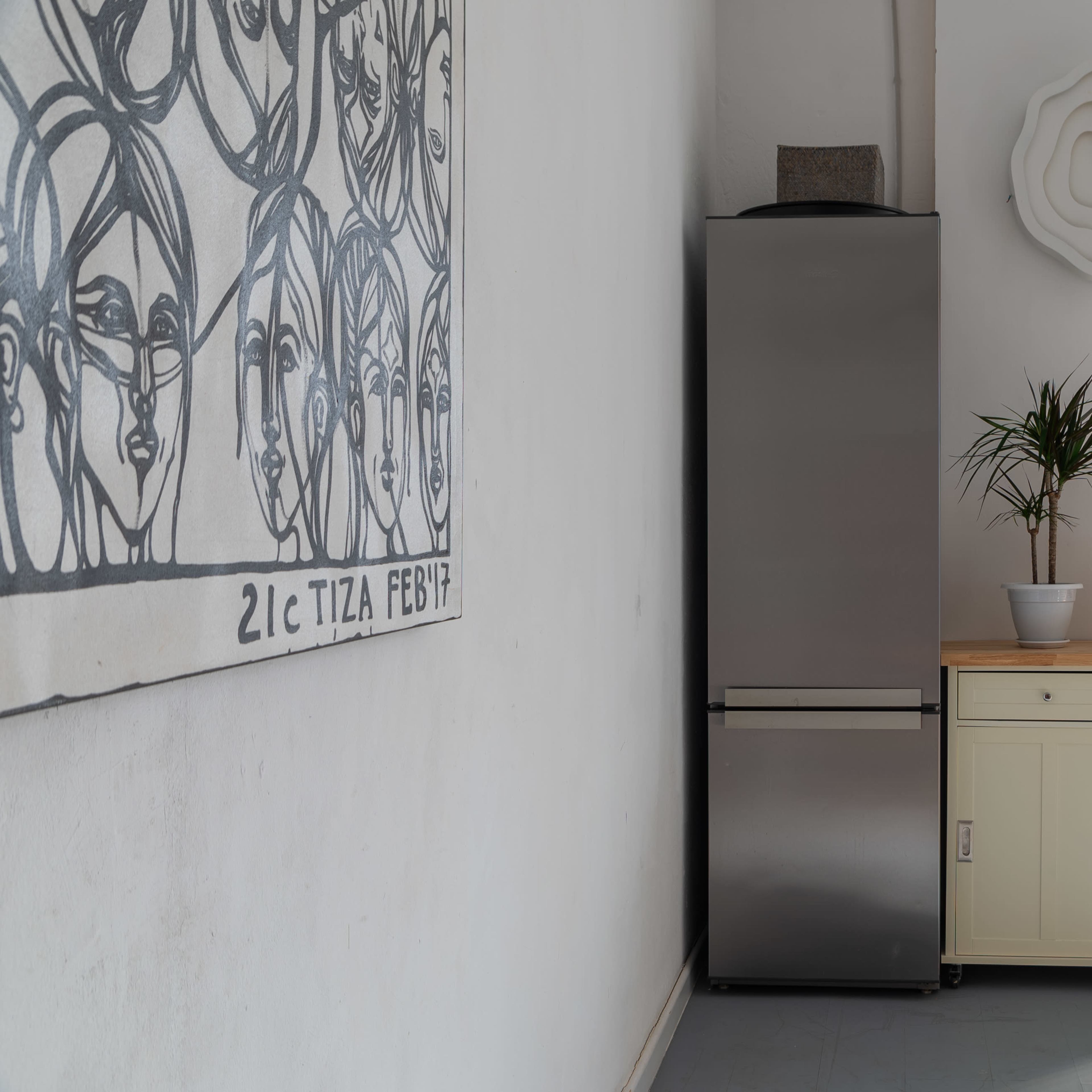 A kitchen corner featuring a silver refrigerator next to a light-colored wooden cabinet with a potted plant and a framed artwork on the wall.
