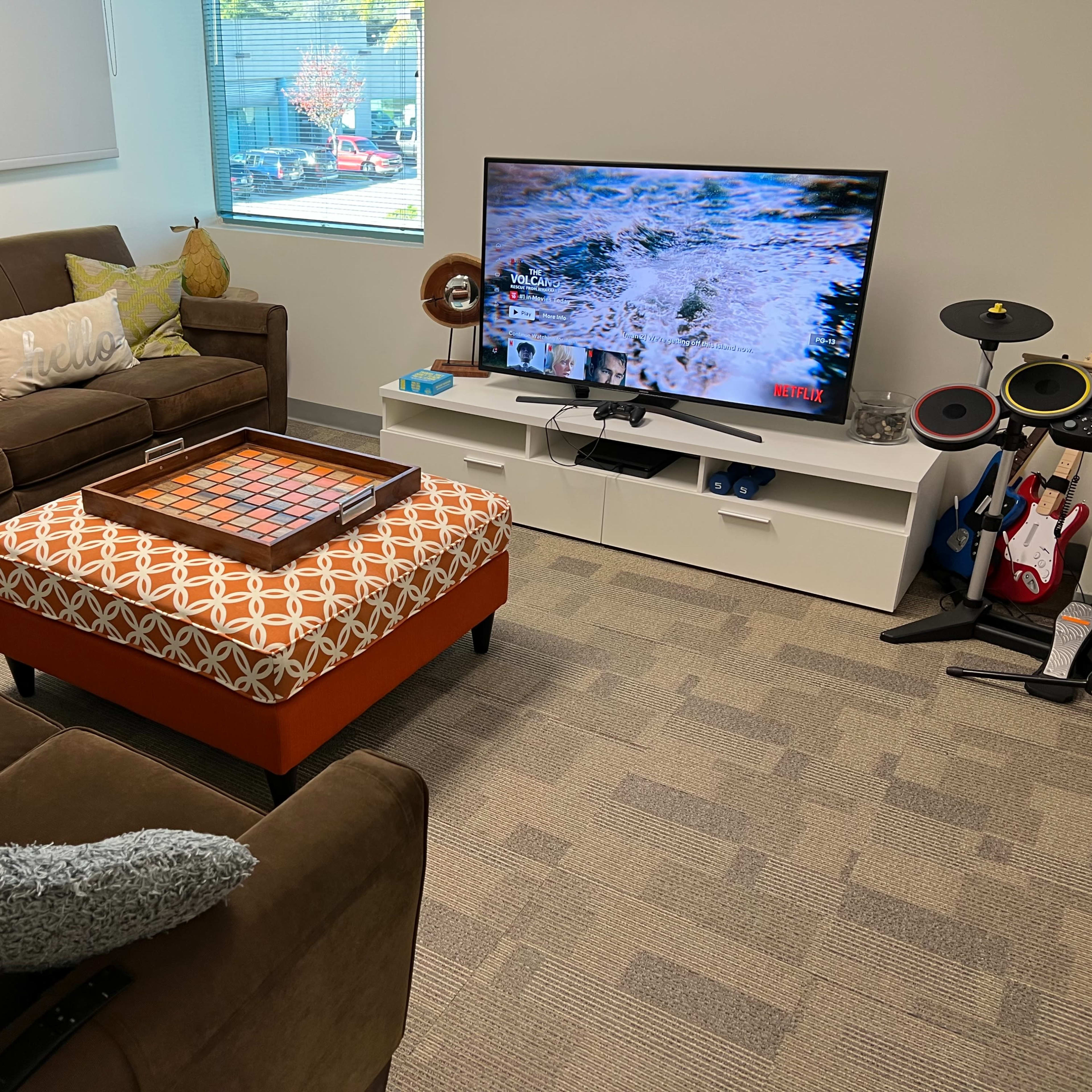 A cozy lounge area with a brown sofa set, a patterned ottoman, and a television on a white media console displaying content from Netflix.