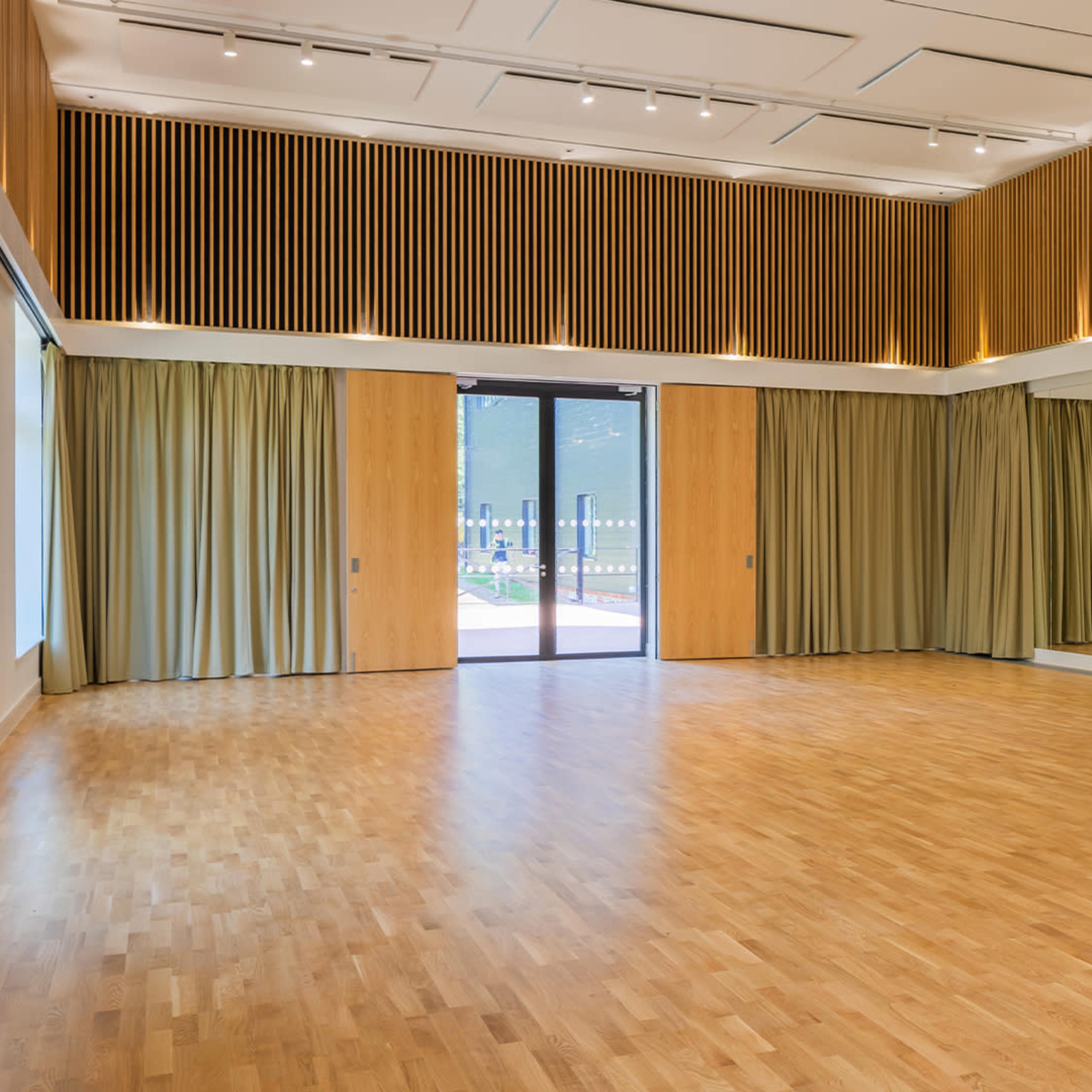 A spacious, empty interior room with wooden flooring, large windows, and curtains, along with a floor-to-ceiling mirror on one wall.