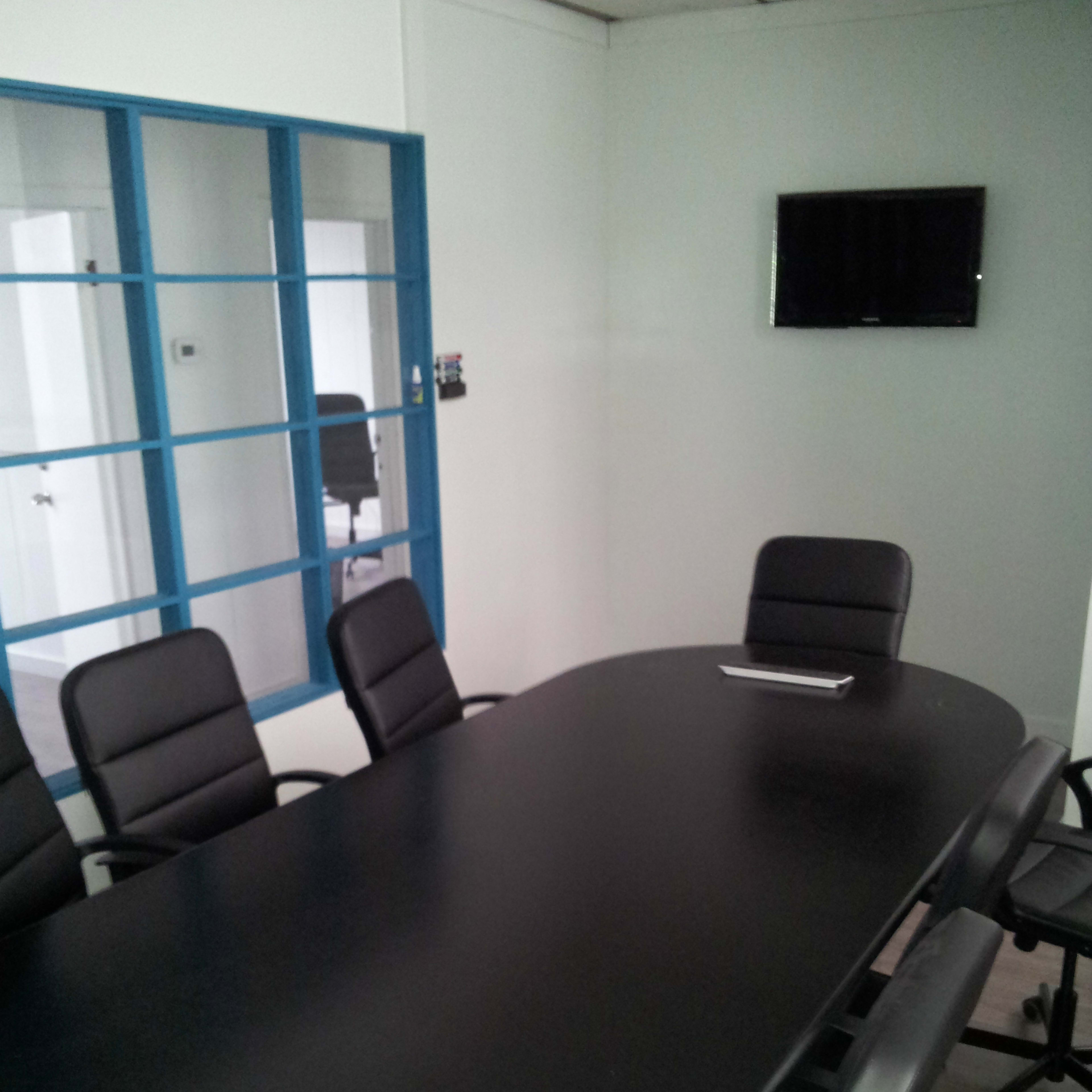 A conference room with a long black table surrounded by black rolling chairs, featuring a wall-mounted TV and a glass-paneled door.