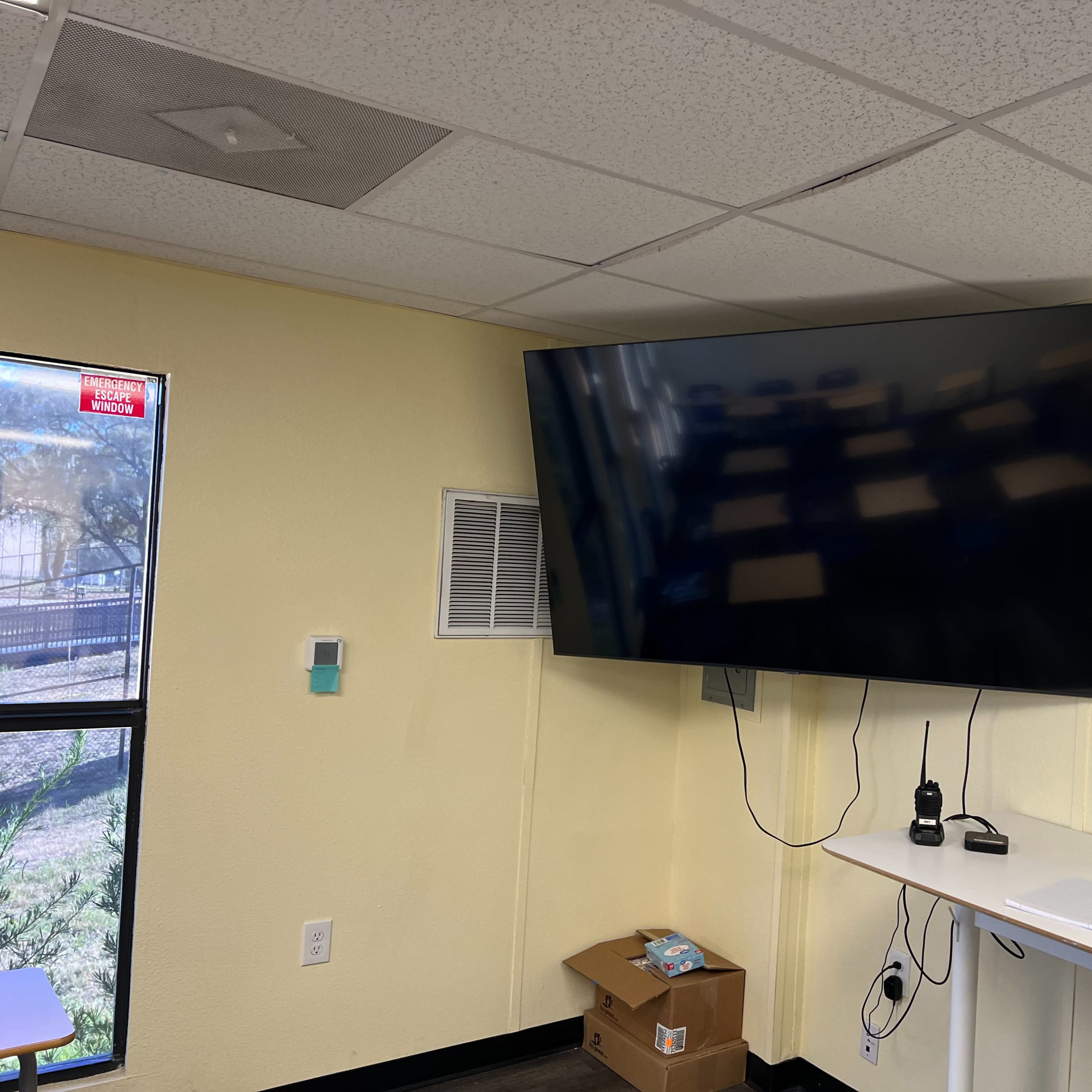 A large television is mounted on a yellow wall in a room, with a window and a table underneath it.