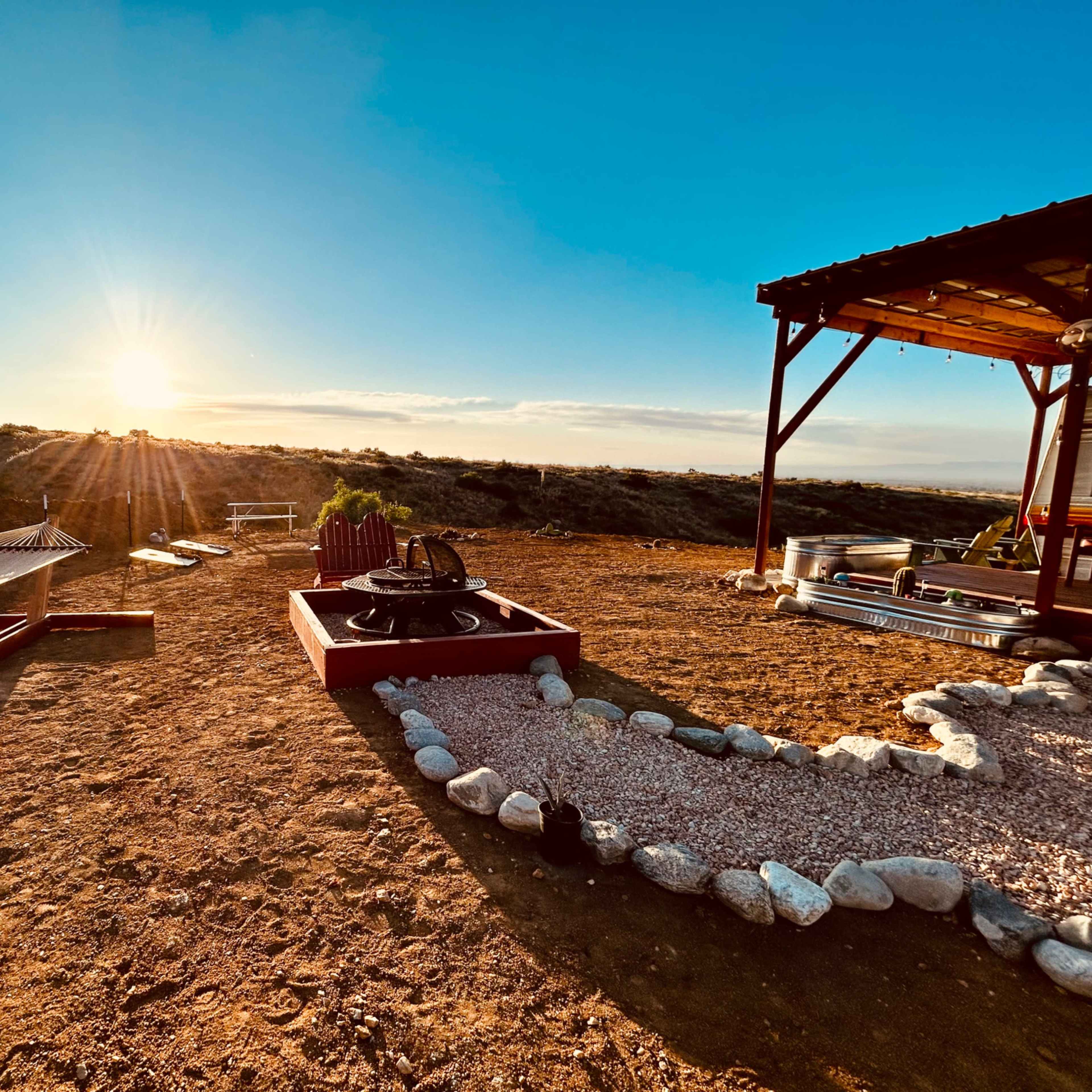 A sunset view with a hammock, a fire pit surrounded by rocks, and a covered patio in a desert landscape.