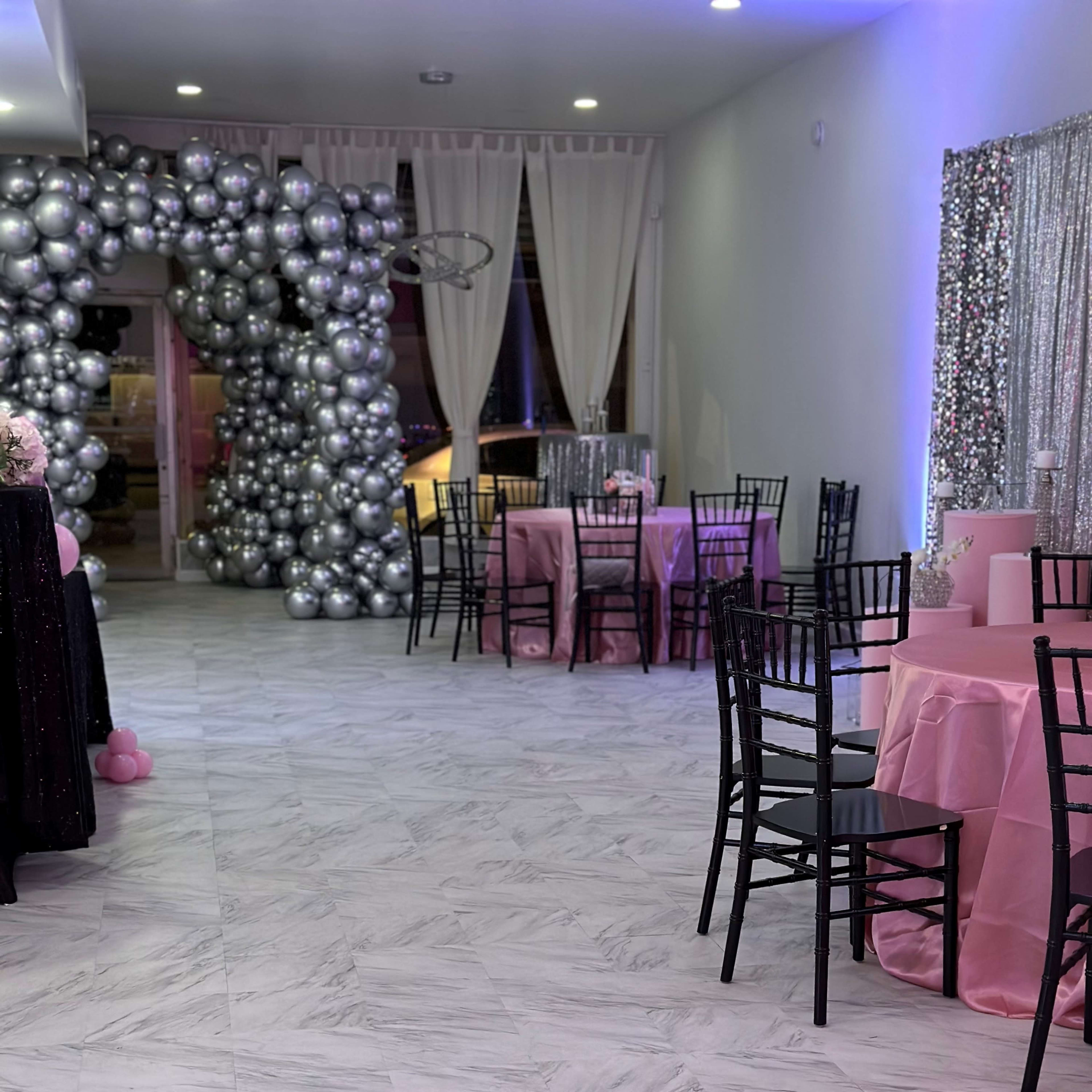 A decorated event space with black chairs and pink tables, accented by a large balloon arch and shimmering curtains.
