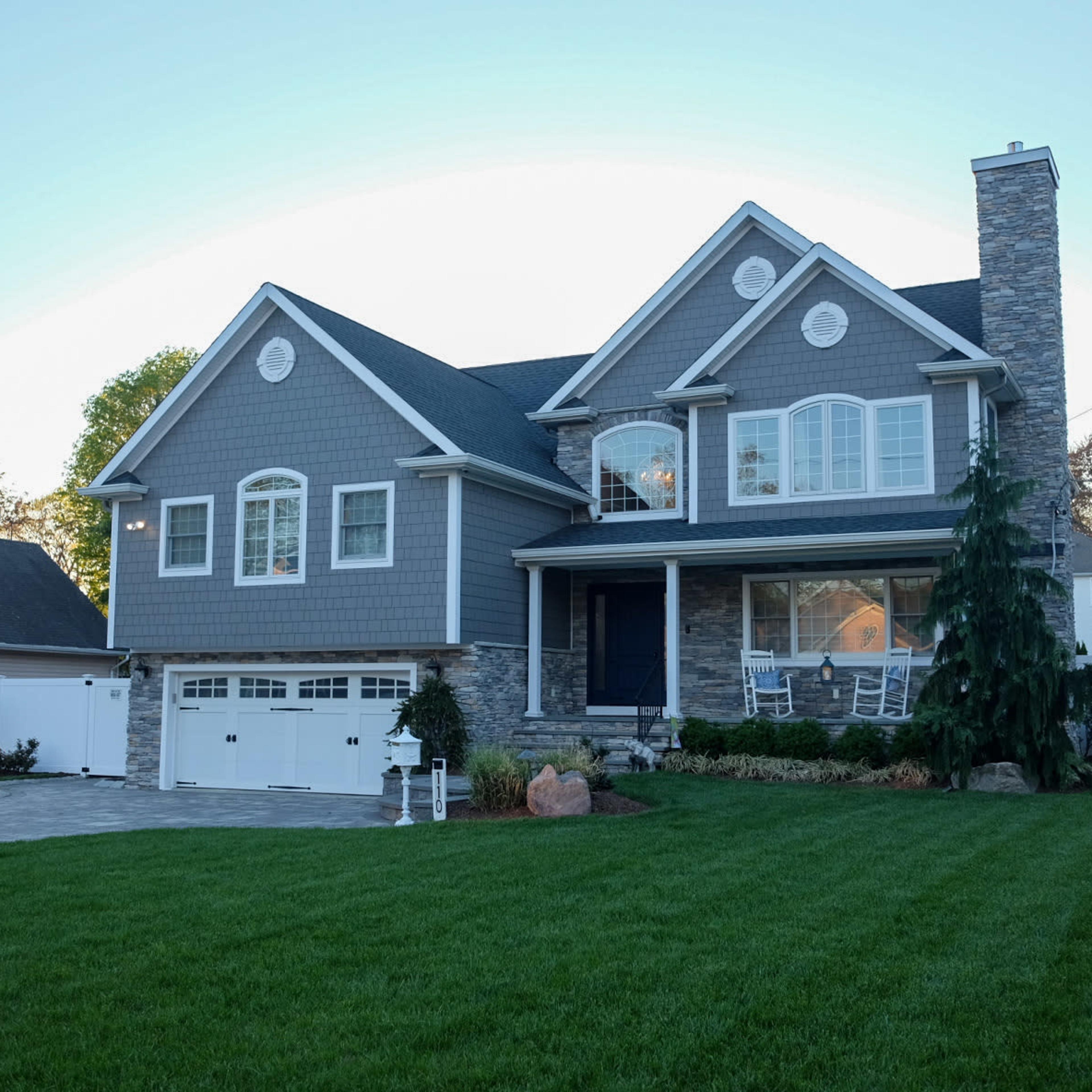 A two-story gray house with stone accents and a front porch, flanked by a neatly landscaped yard and an American flag.
