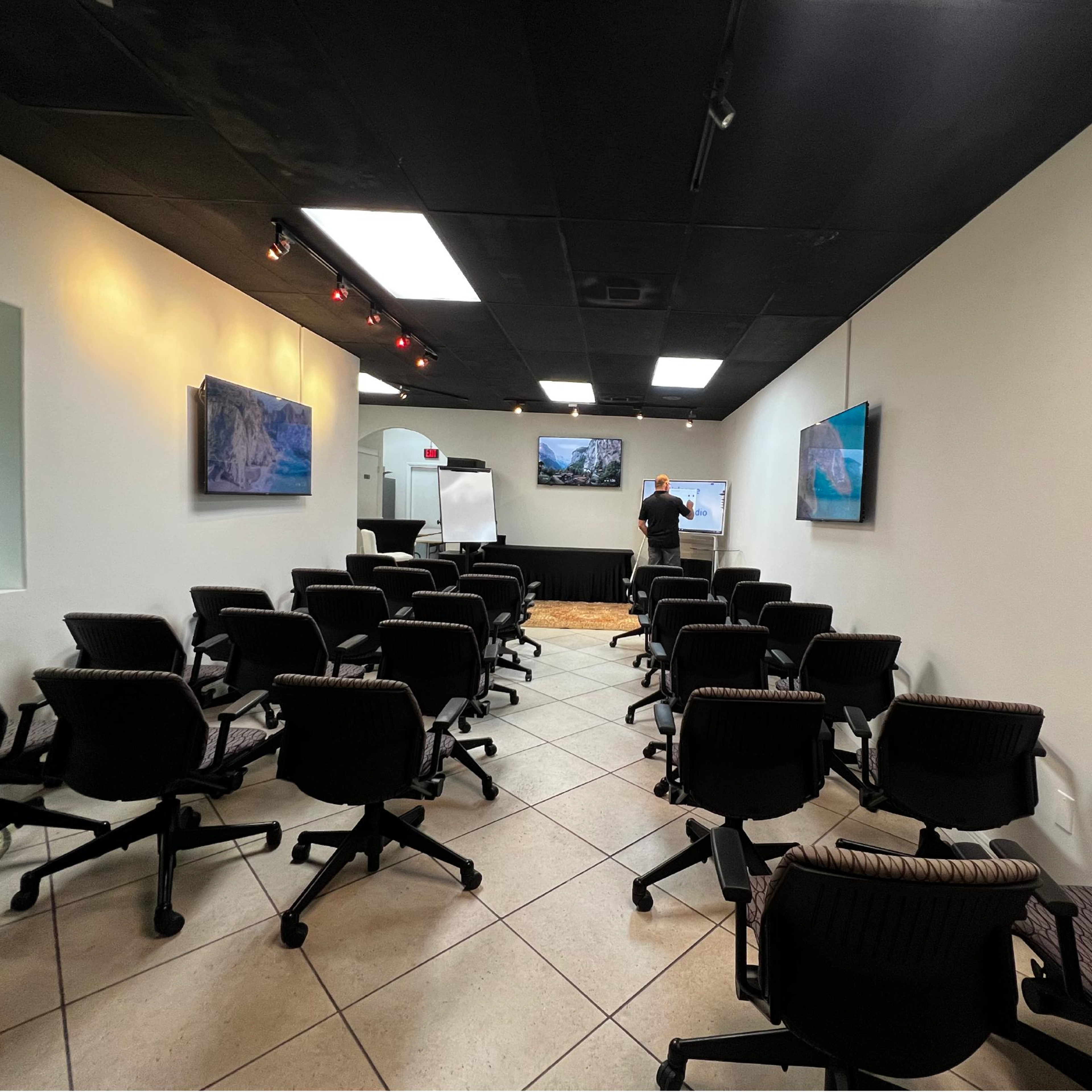 A man stands in a conference room filled with rows of empty, swivel chairs facing whiteboards and screens.
