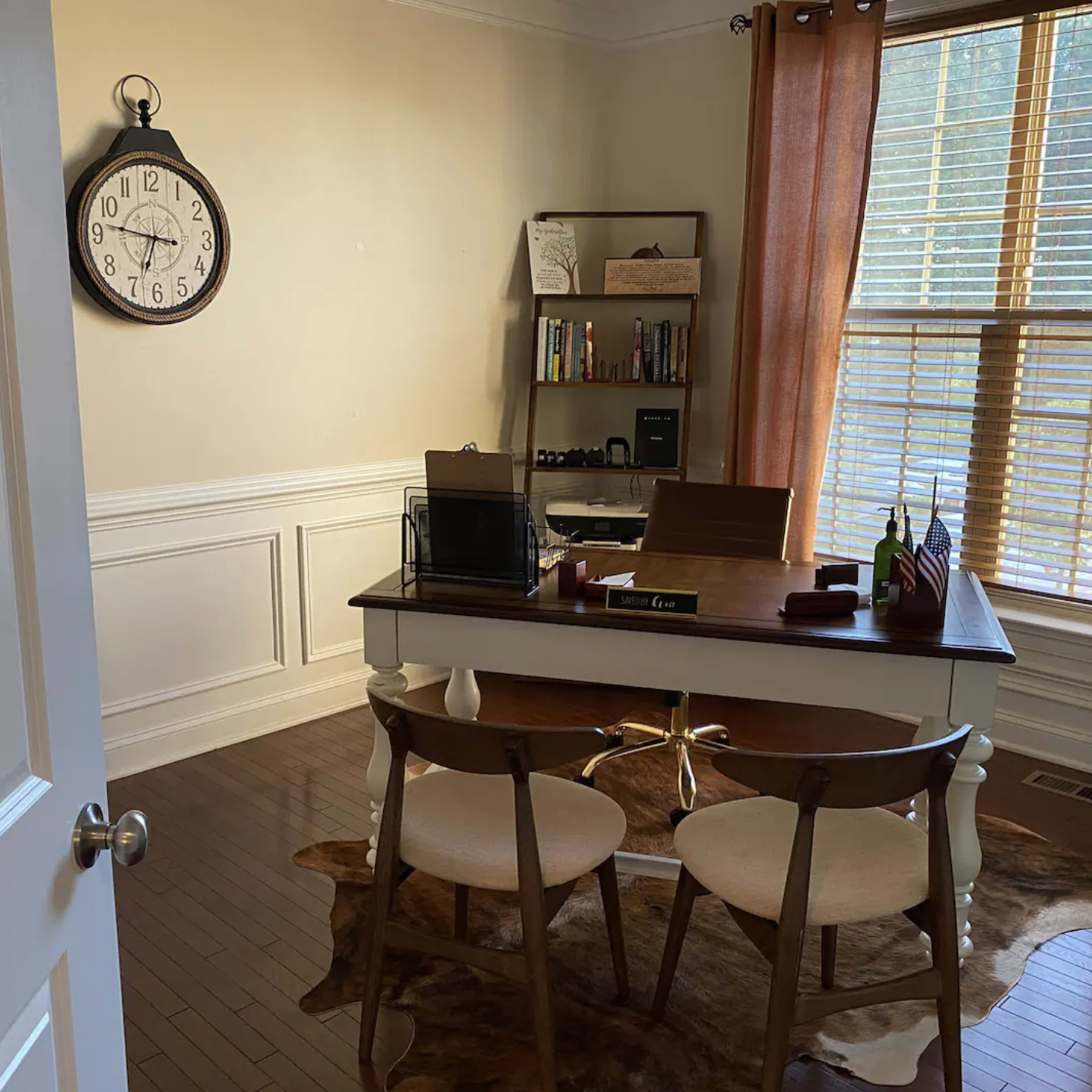 A well-lit home office features a wooden desk with two chairs, a clock on the wall, and shelves filled with books.
