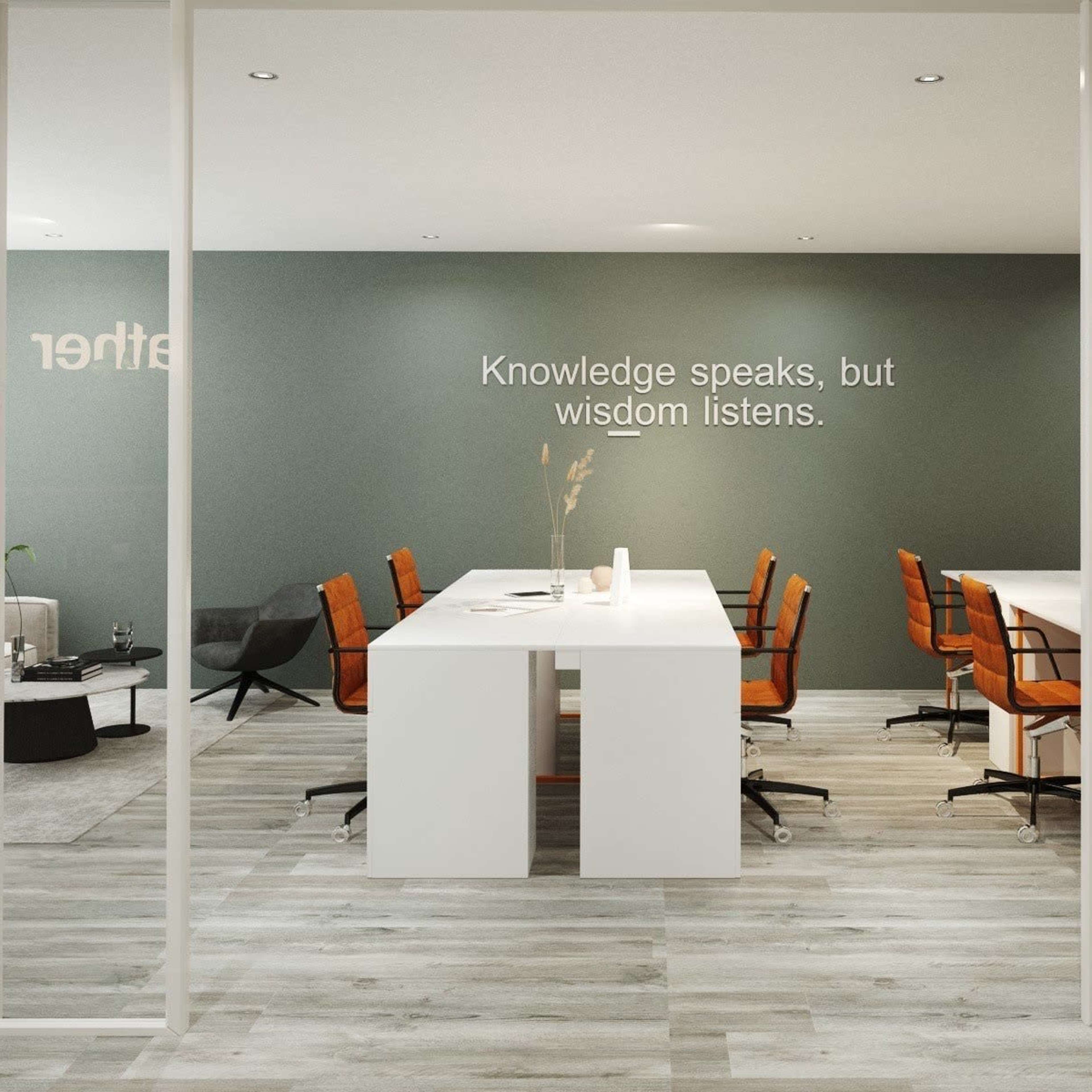 A modern office space features a large white table surrounded by orange chairs, with a quote on the wall and a lounge area in the background.