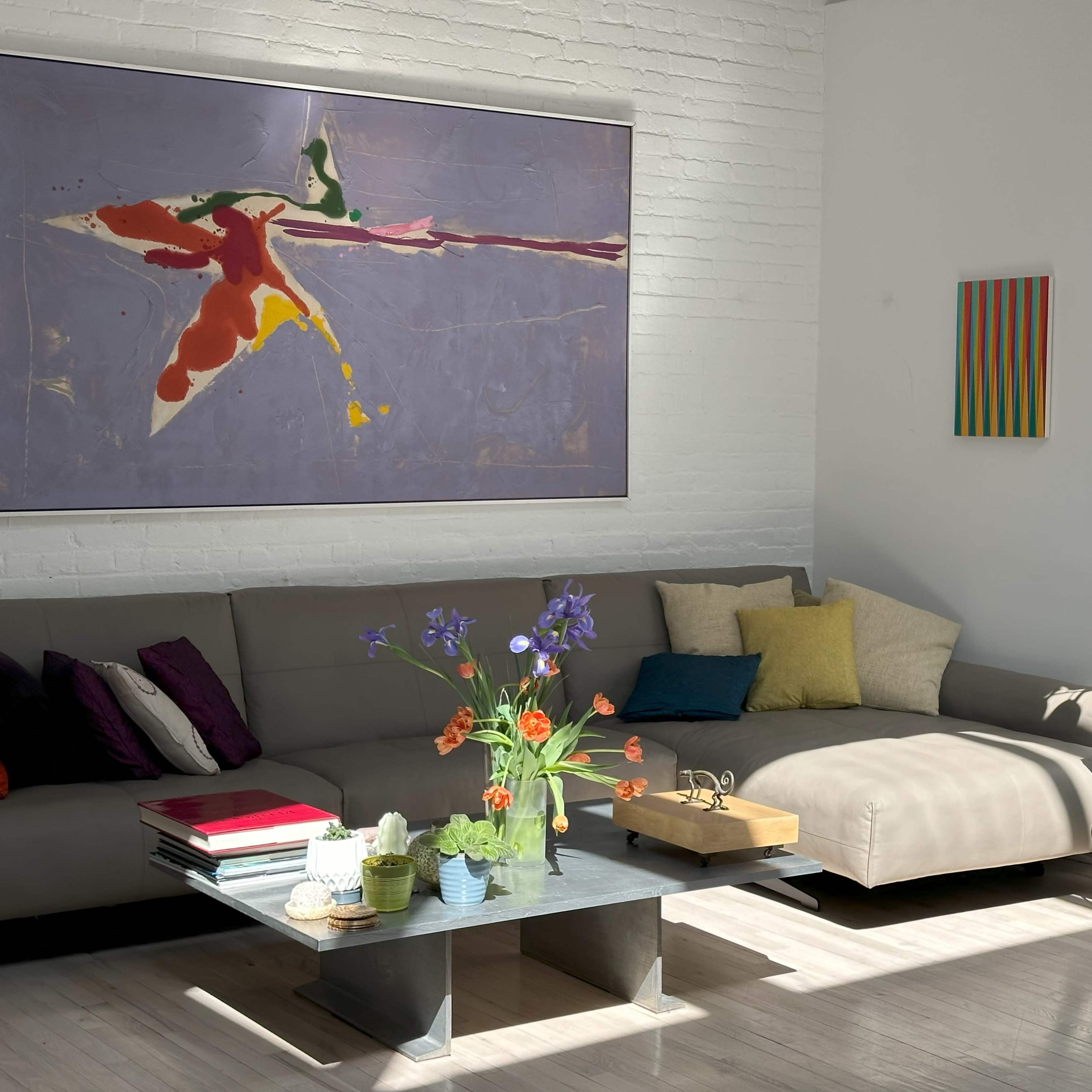 A sunlit living room features a gray sofa adorned with colorful pillows, a coffee table with books and plants, and vibrant artwork on the walls.