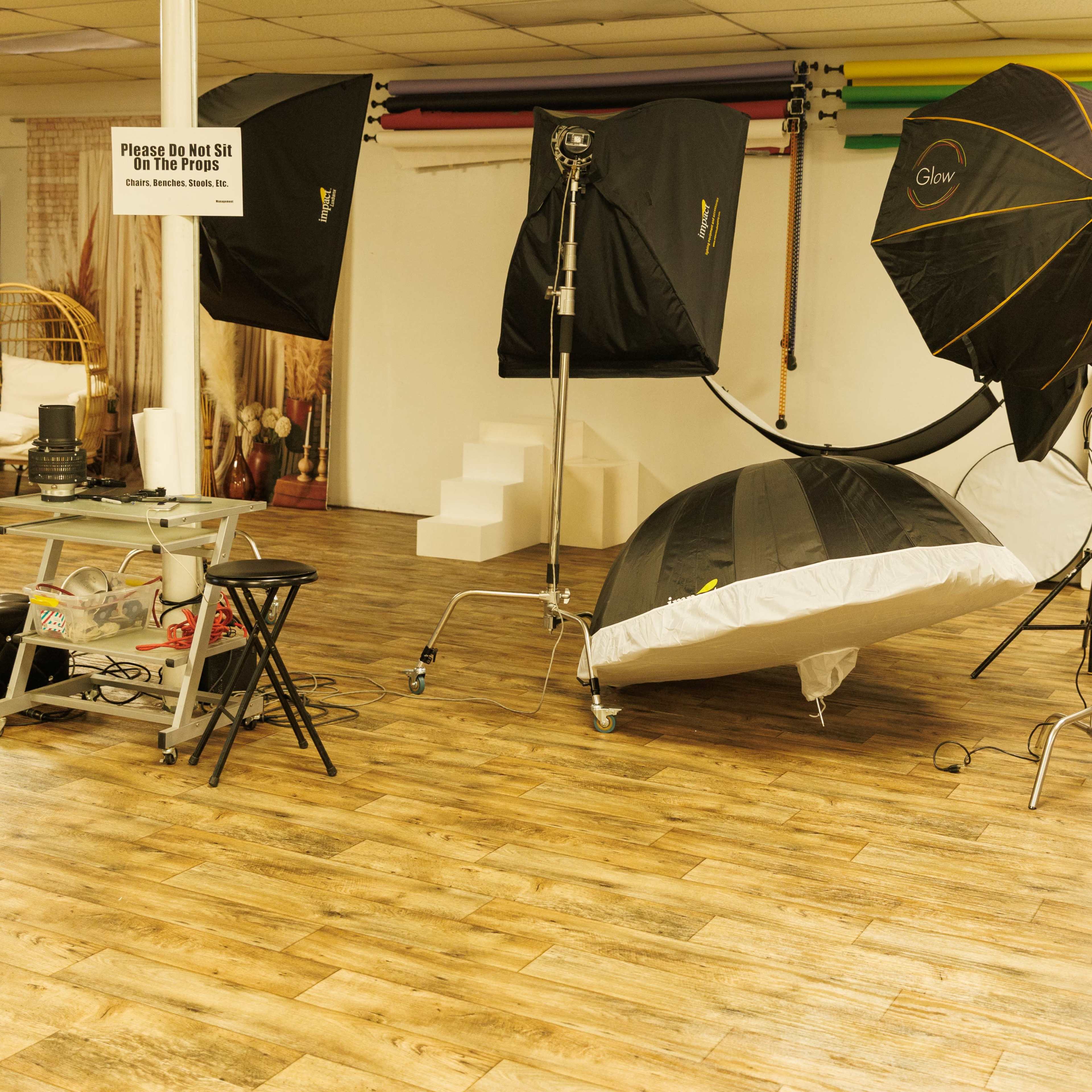 A photography studio setup with multiple softbox lights, a large reflective umbrella, and a low-profile bed on a wood-finish floor.