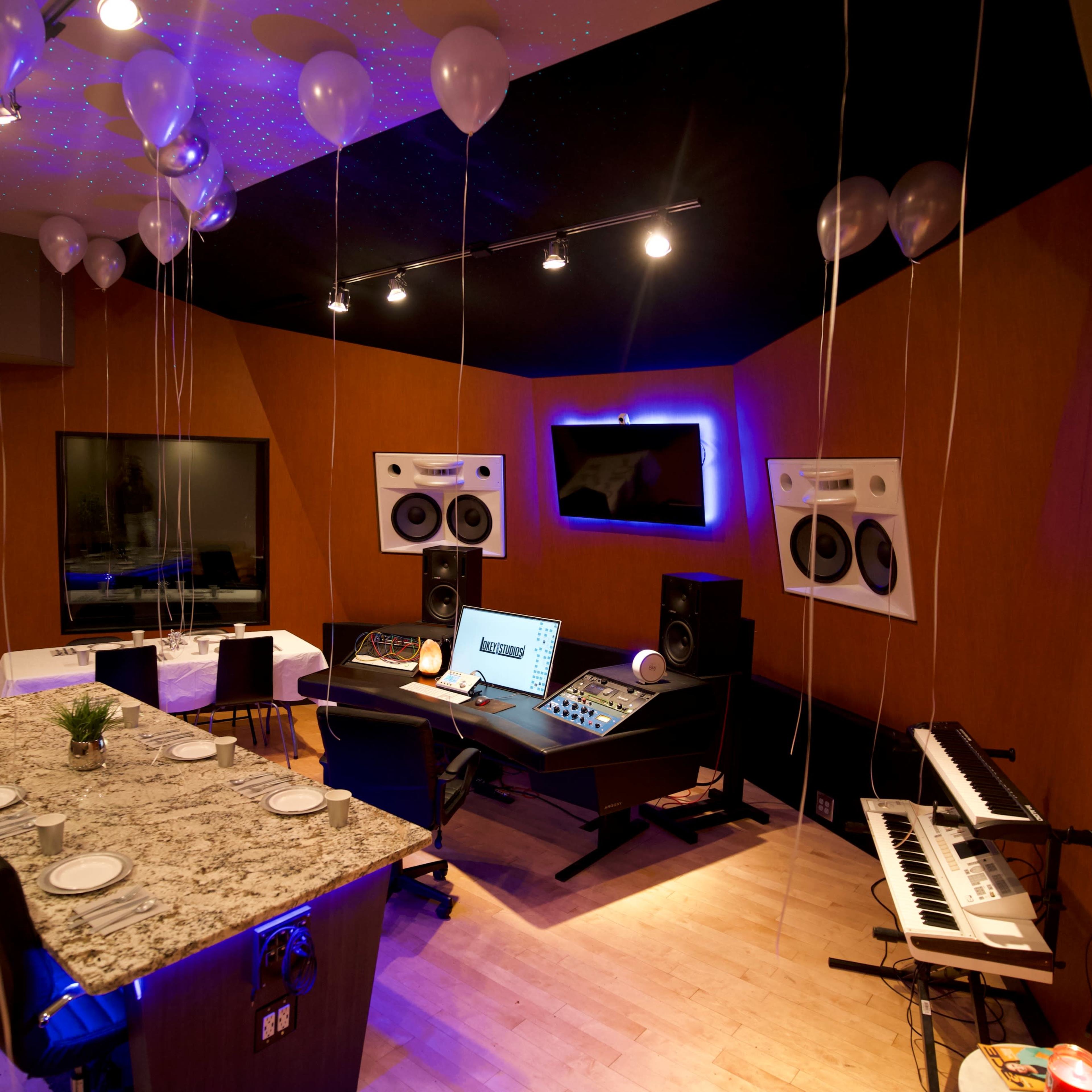 A modern music studio featuring a central mixing console, audio speakers, a keyboard, and a dining table surrounded by balloons and softly illuminated ceiling lights.