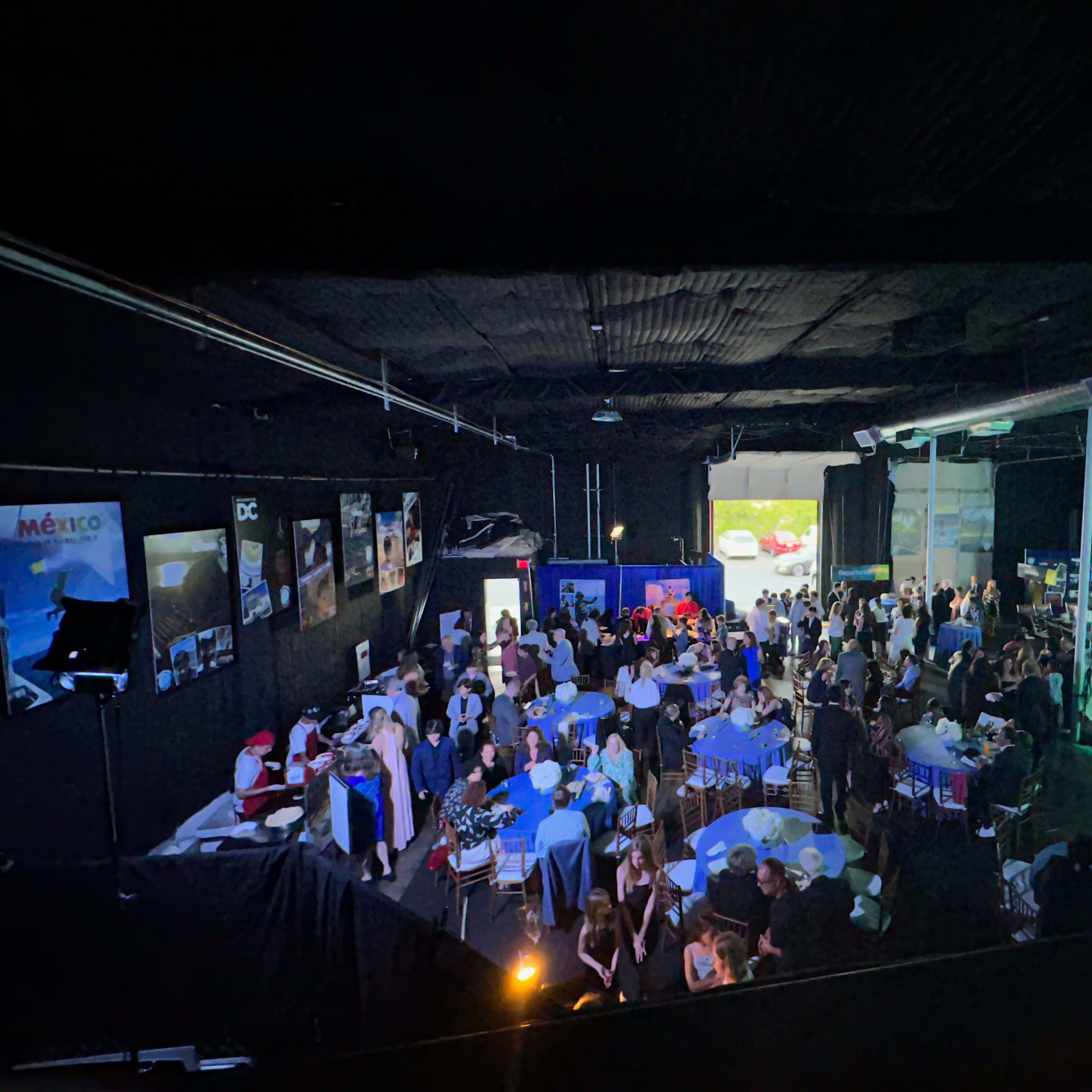 A large gathering of people is taking place inside a dark, spacious venue, featuring tables with blue tablecloths and decorations, while a stage and screens are set up in the background.