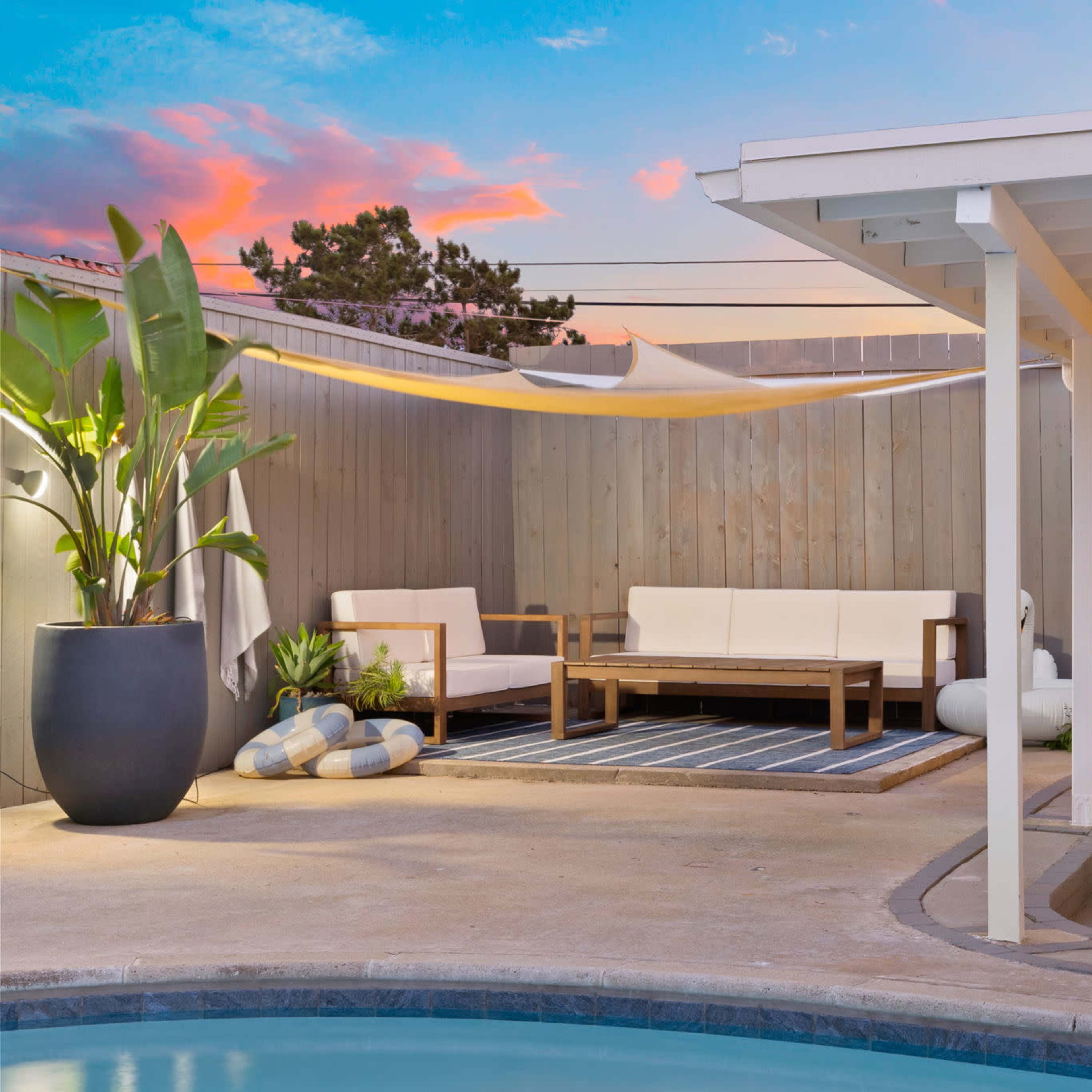 The scene features a modern outdoor living area with a pool, comfortable seating, and a dining space, surrounded by wooden fencing and plants under a colorful sunset sky.