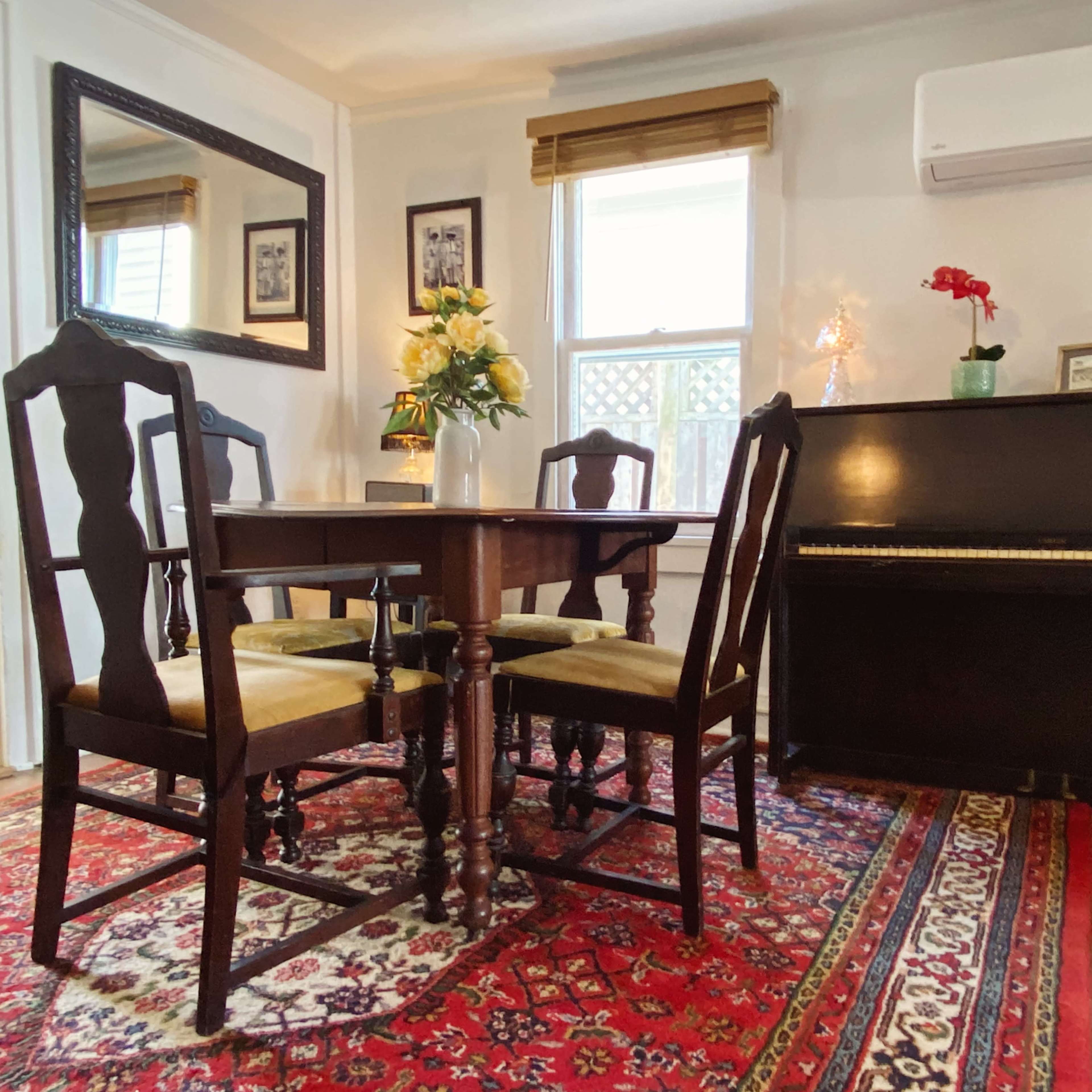 A dining area features a wooden table with four chairs, a piano, and a rug with intricate patterns.