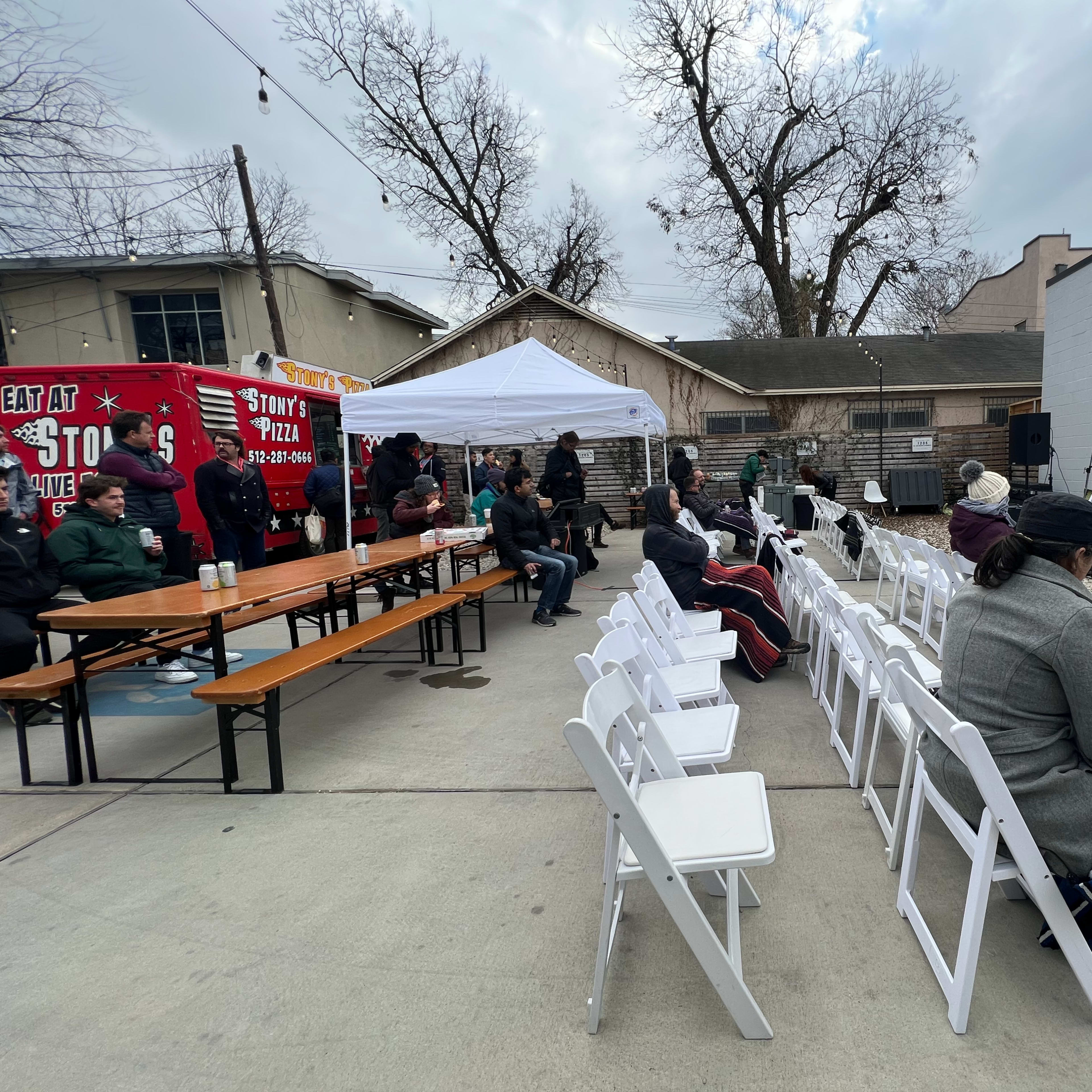 A crowd of people sits in folding chairs arranged in front of a small stage with a projector screen, while a food truck is parked nearby in a shaded outdoor area.