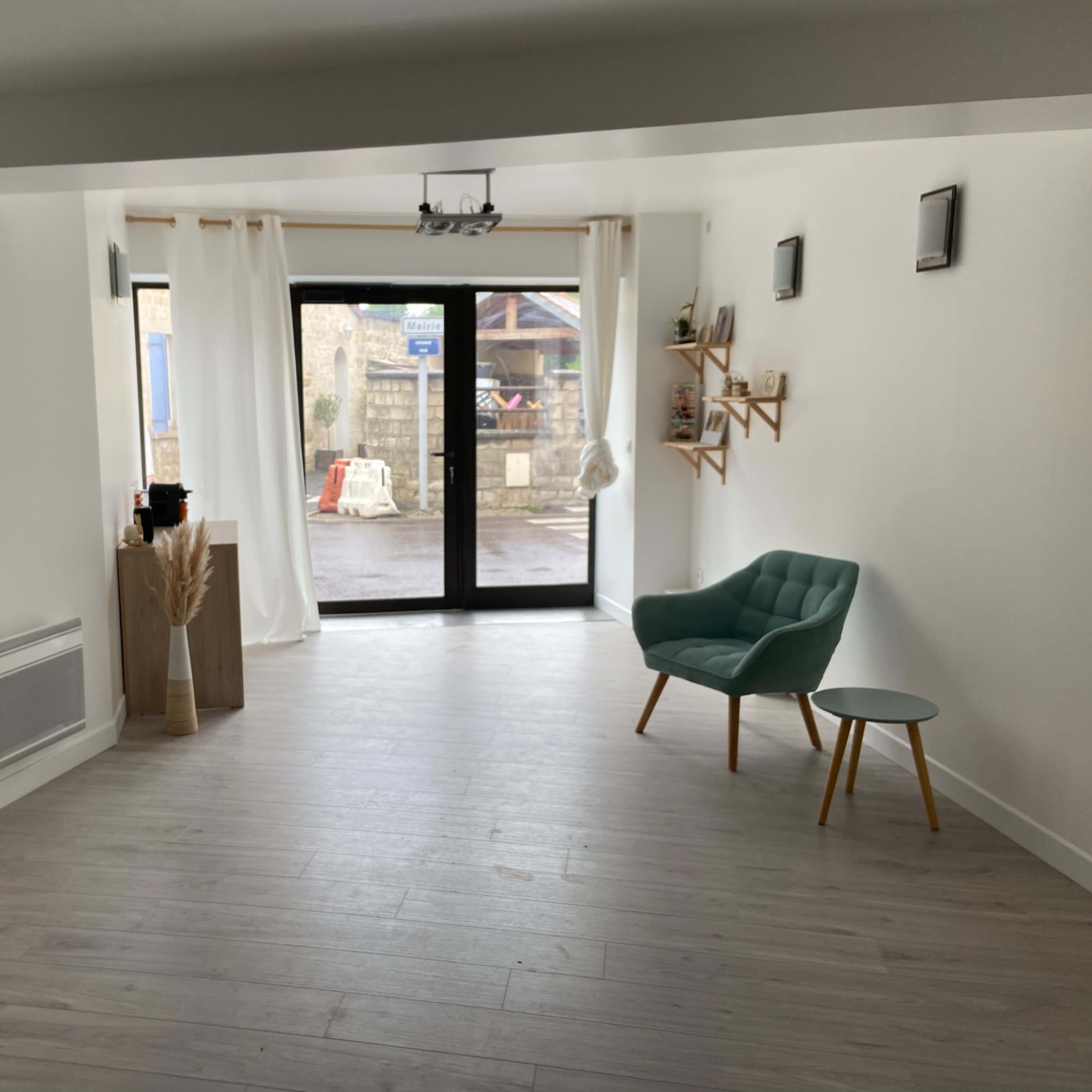 The interior of a minimalist shop features a green chair and small table, a clothing rack with garments, and large windows facing a street.