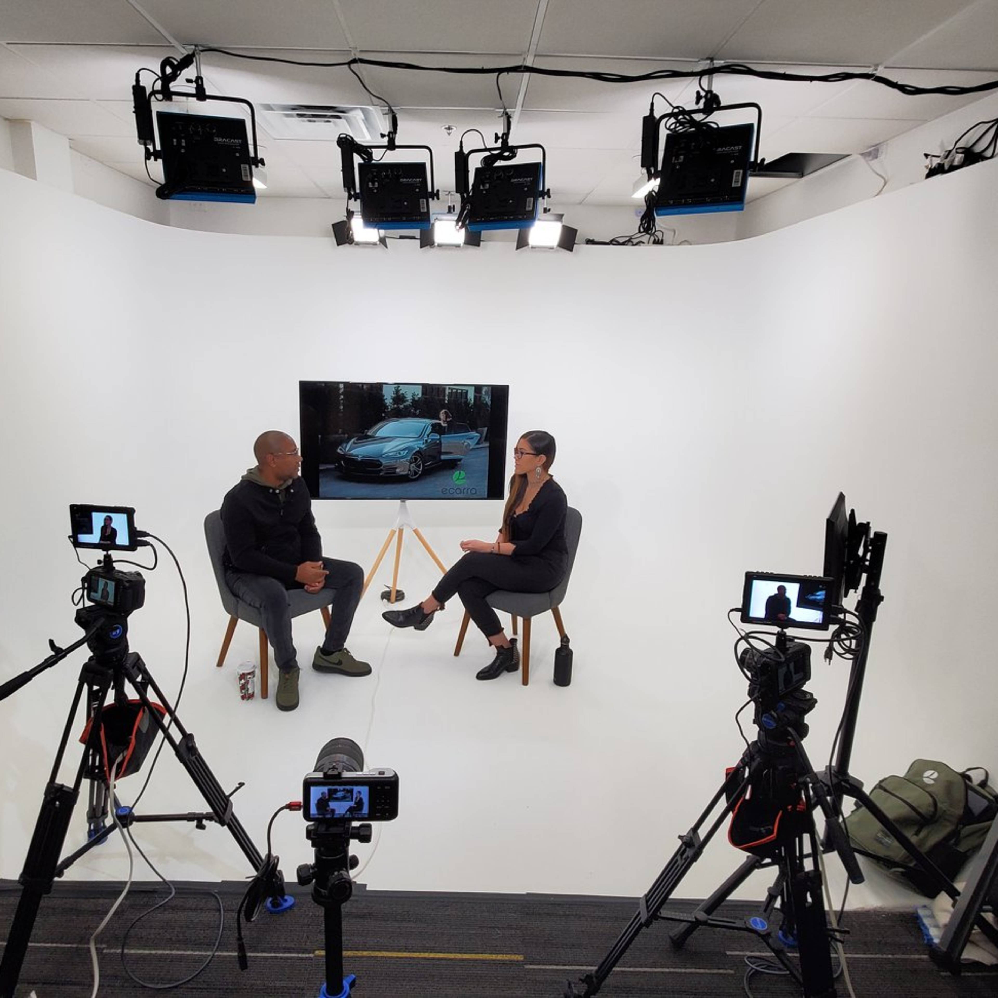 A man and a woman sit across from each other in a studio setting with multiple cameras and bright lights, while a large screen displays an image of a car.
