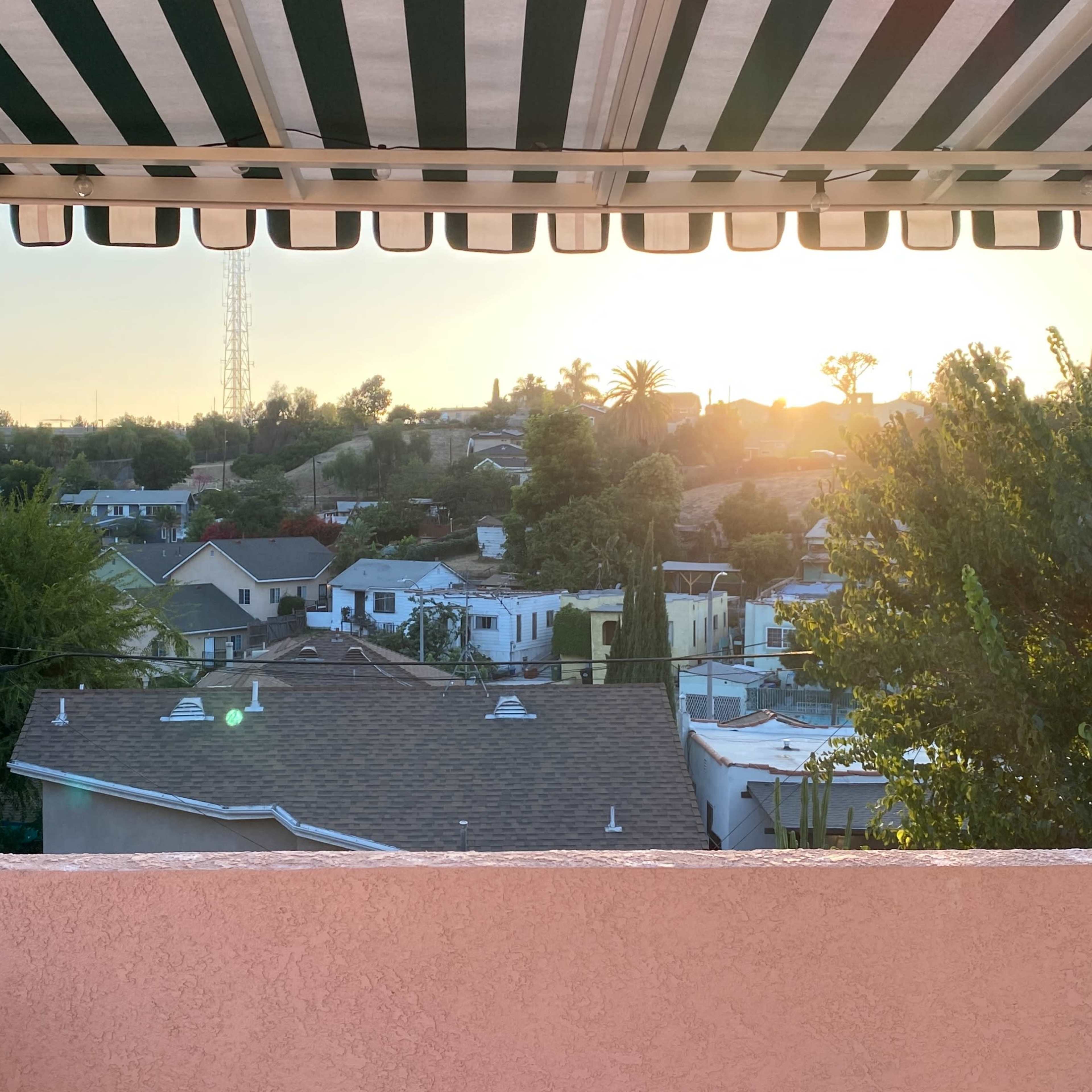 A view from a balcony featuring a sunset over residential houses and trees, framed by a striped awning.