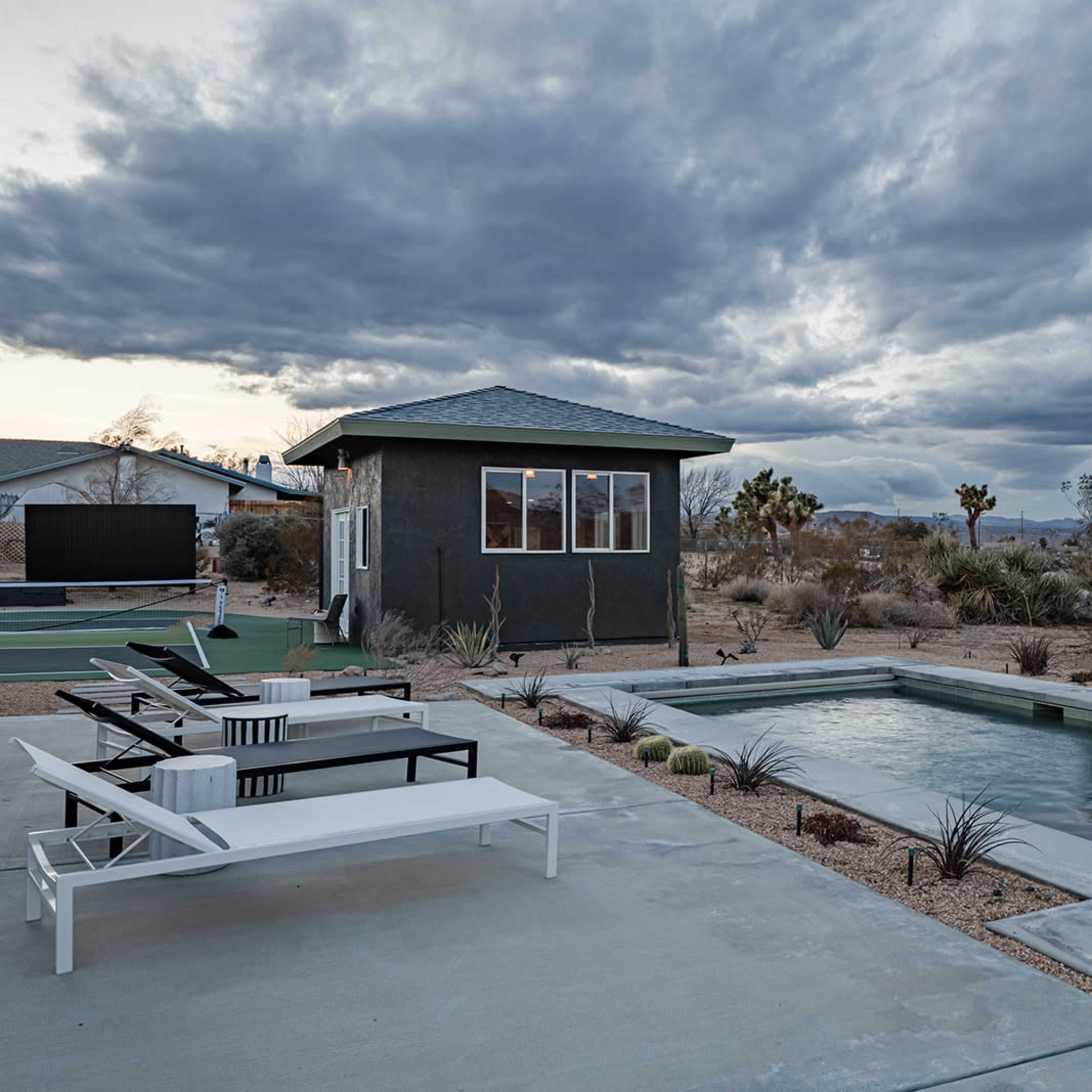 A swimming pool surrounded by desert landscaping and lounge chairs is situated next to a small building under a cloudy sky.