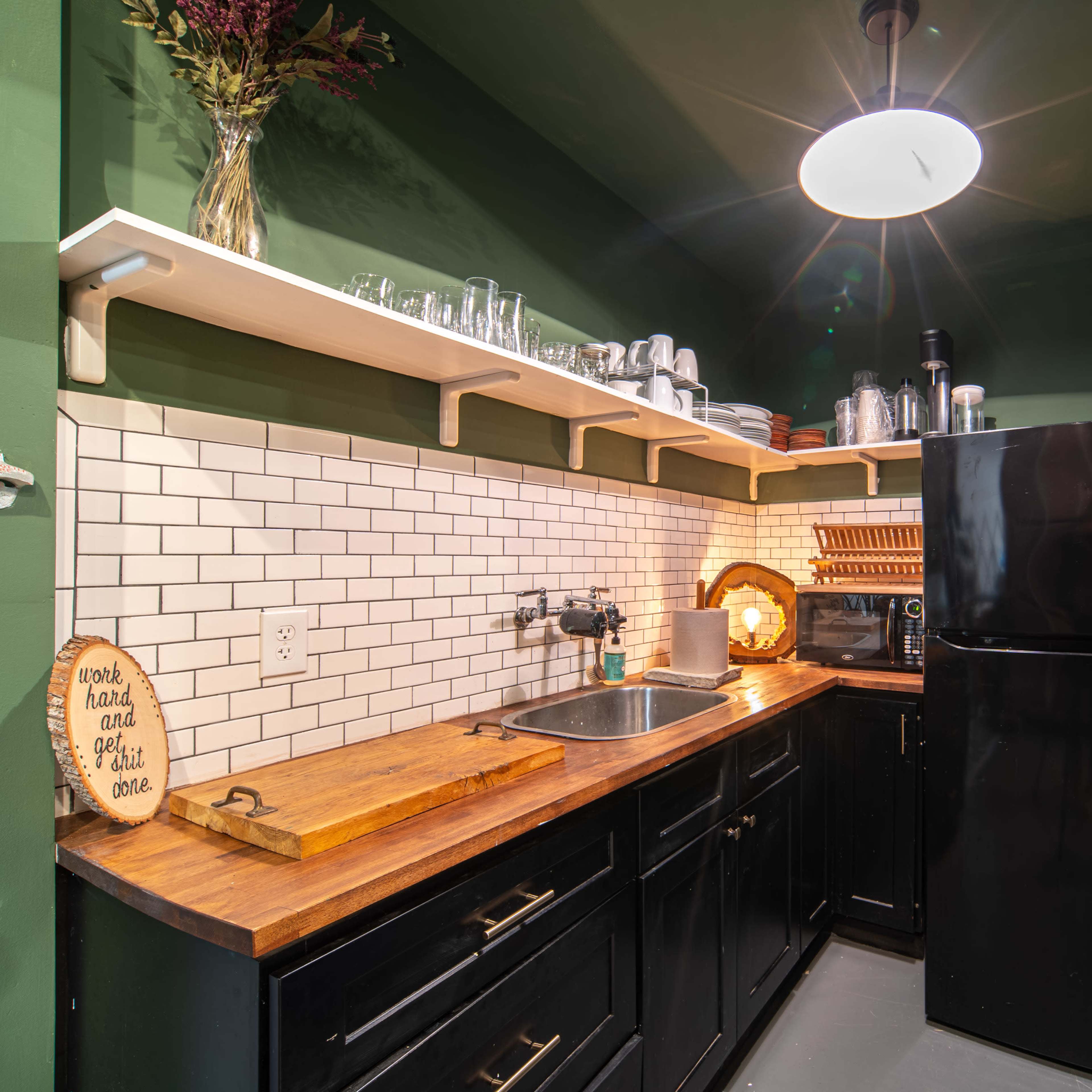 A small kitchen area featuring a black refrigerator, a stainless steel sink, wooden countertop, open shelves with glassware, and a tiled backsplash, all set against green walls.