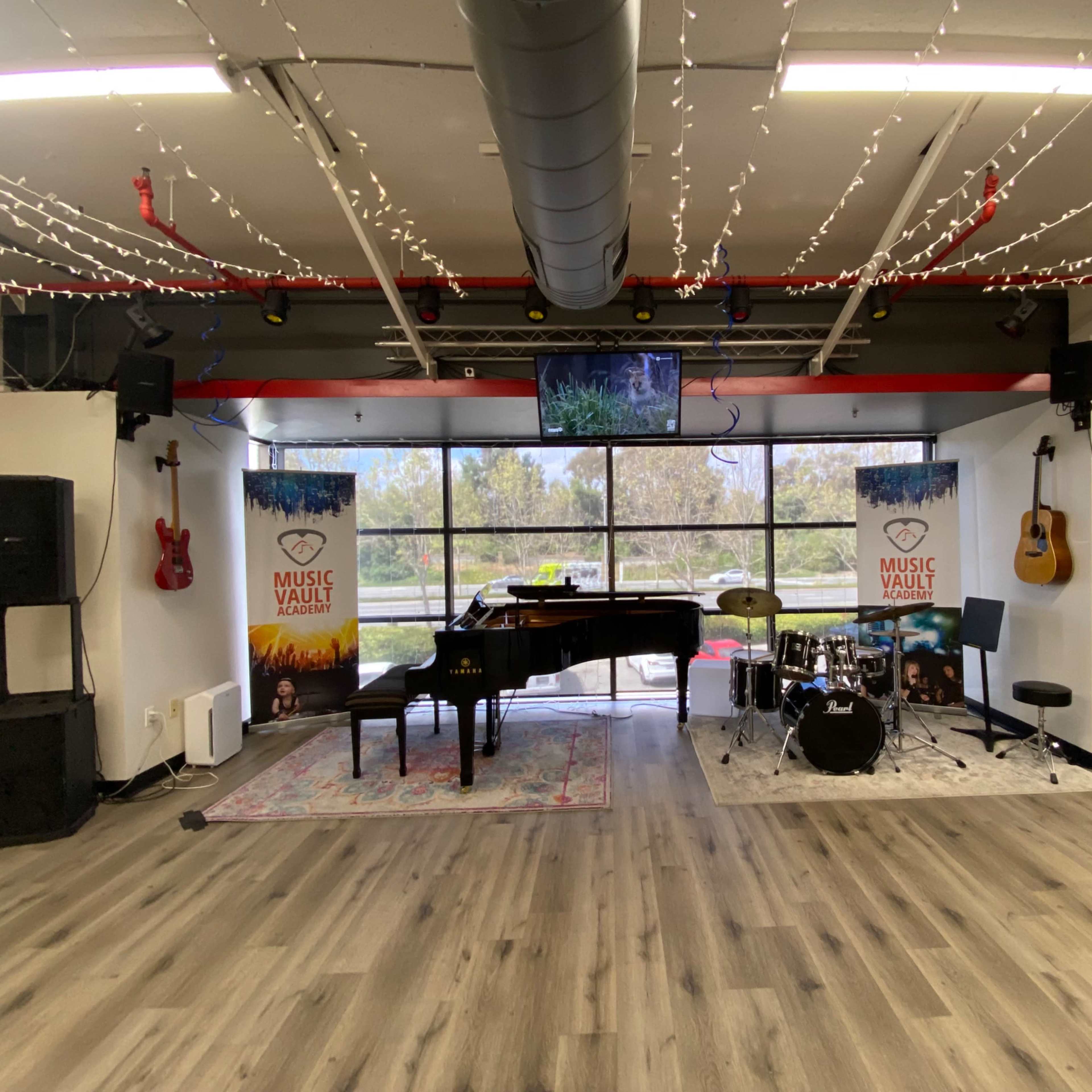 A music studio setup featuring a grand piano, a drum set, and electric guitars flanked by banners for "Music Vault," with string lights overhead and large windows providing natural light.